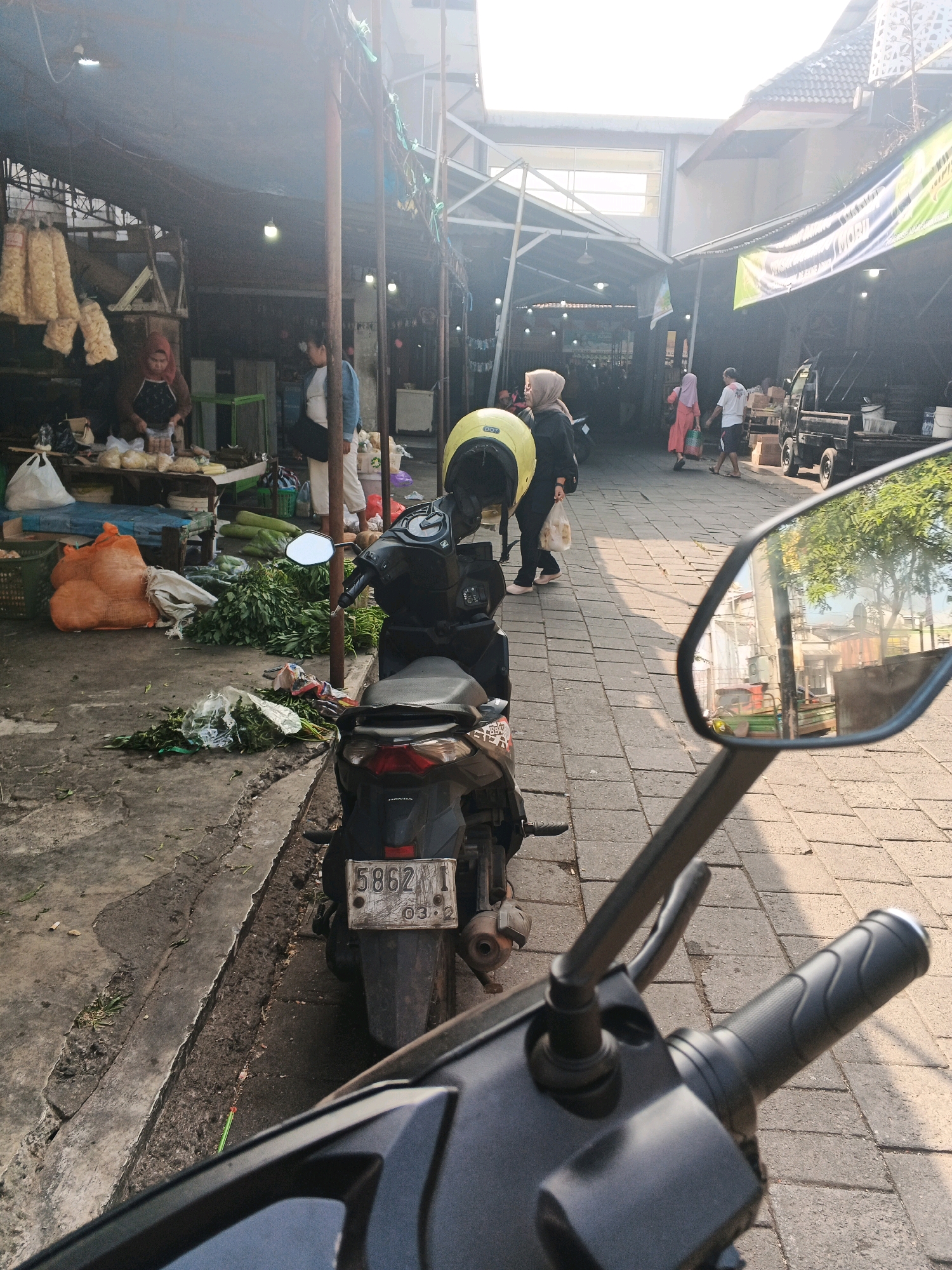 The image depicts a street scene in what appears to be a market in Ungaran, Indonesia. In the foreground, the handlebars and side mirror of a black motorcycle are visible. The side mirror reflects trees and buildings. Behind the motorcycle, a market stall is set up with various goods, including large orange sacks and bundles of green vegetables. Several people are present, some appearing to be vendors and others customers. A woman in a headscarf is seen arranging items at a stall, while other individuals walk along the paved walkway. The environment is characterized by stalls with tarpaulin roofs, a paved walkway, and natural lighting suggesting it is daytime. There is a general atmosphere of a bustling local market.