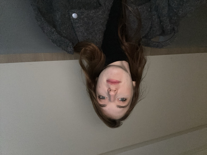 A young adult female with fair skin, long reddish-brown hair, and blue eyes is depicted from the chest up, looking directly at the camera with a neutral expression. She wears a black turtleneck top and a textured dark grey coat, visible with a silver-colored snap button on the right side of the frame. The background consists of a plain, light-colored wall above a dark grey horizontal surface. The lighting is dim, appearing to be artificial. The specific location within Huangdao District, China, cannot be determined from the visual information provided in the image.