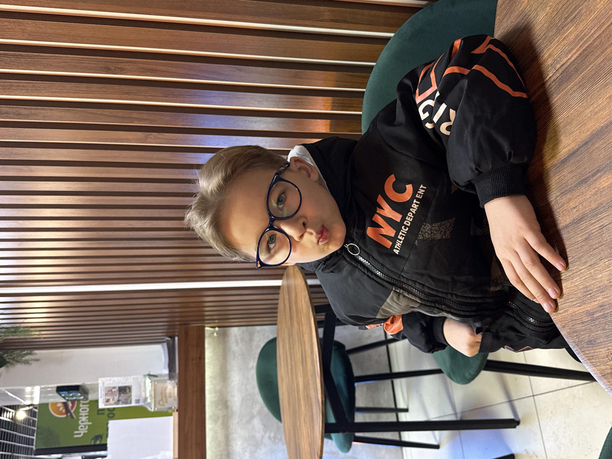 A young person with blond hair and blue glasses is seated at a wooden table in what appears to be a cafe or restaurant. The person is wearing a black jacket with orange accents and the letters "NYC" on the front. They are making a pouting face towards the camera. Behind them, there is a wall with horizontal wooden slats. There are also green chairs and tables in the background. A sign in Russian with "Черного" is visible in the lower-left corner of the image, along with other menu items. The picture appears to have been taken in Rostov-on-Don, Russia.