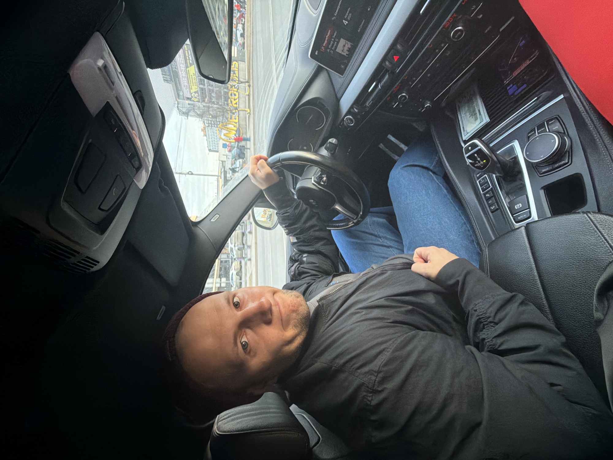 A man is sitting in the driver's seat of a car in Krasnoyarsk, Russia. The car is stopped on a street with buildings and signage visible through the windshield. The man is holding the steering wheel with his left hand and looking at the camera with a quizzical expression. He is wearing a dark jacket and blue jeans. The interior of the car is visible, including the dashboard, gear shift, and center console. A stack of bills can be seen on the center console.