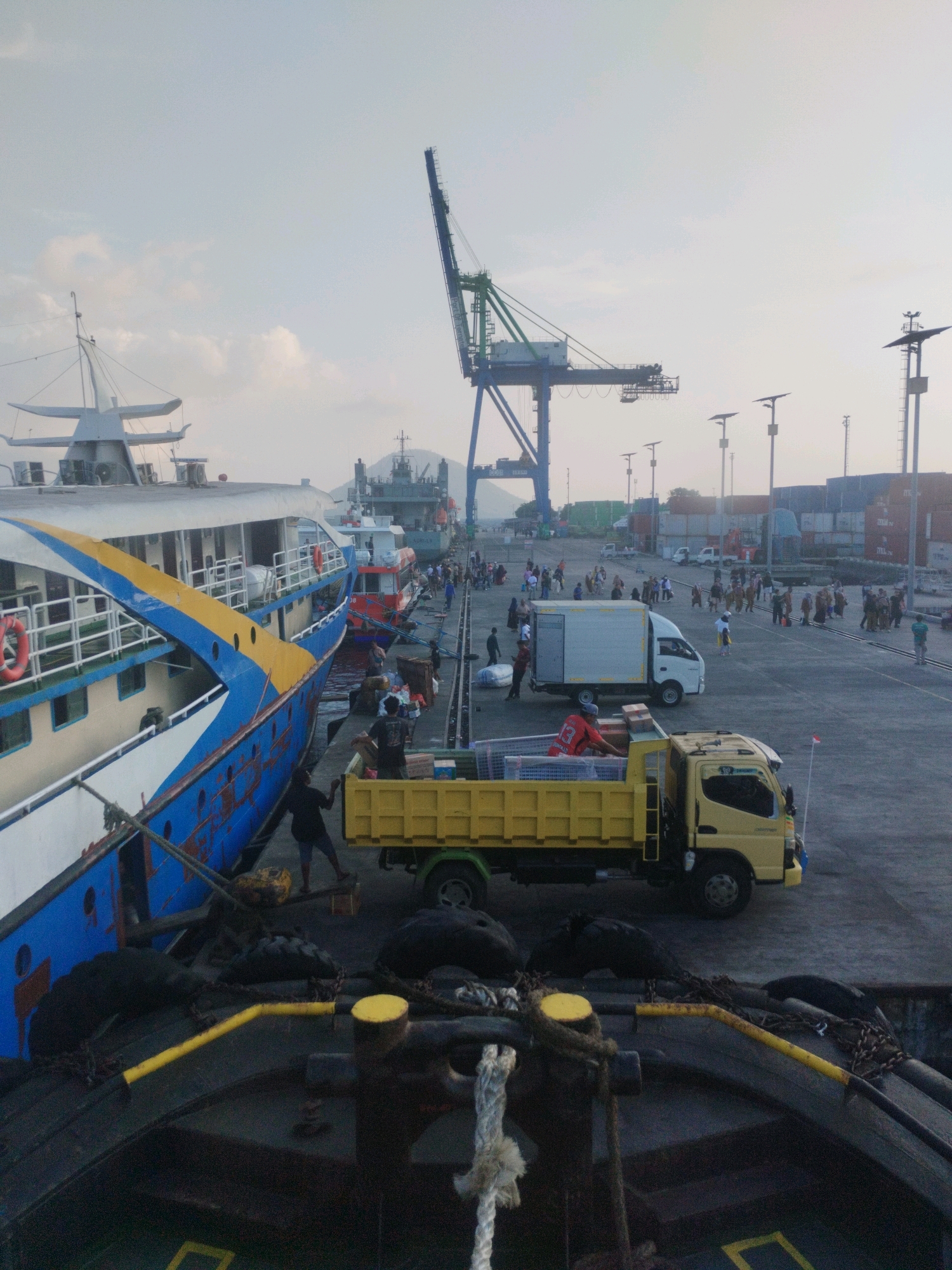 The image shows a busy port scene at Ternate, Indonesia. In the foreground, a large blue and yellow ferry is docked. Several people are visible on the pier, some engaged in loading or unloading cargo. A yellow dump truck is parked nearby, and a white box truck is also present. In the background, a large gantry crane stands tall, indicative of a cargo port. Shipping containers are stacked in the distance. The sky is overcast, suggesting it might be early morning or late afternoon. The overall impression is one of a functional and active maritime hub.