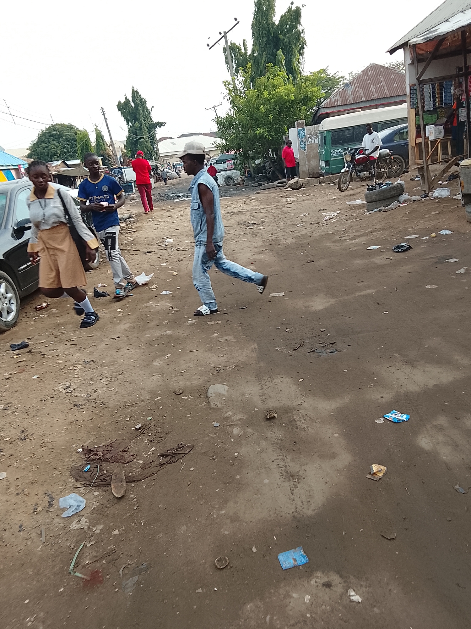 The image shows a dirt road in what appears to be a commercial area. Several people are present: a young woman in a light-colored top and khaki skirt walks near the left edge of the frame; a young man in a blue soccer jersey and jeans walks behind her; a man in a denim outfit and a white hat walks across the road. Further back, a man in red clothing walks away. In the background, a man sits on a motorcycle near a shop. The road is littered with trash. A car is parked on the left side of the frame. Buildings and trees are visible in the background. Based on the available information, it's not possible to confirm if this photo was taken in Abuja, Nigeria.