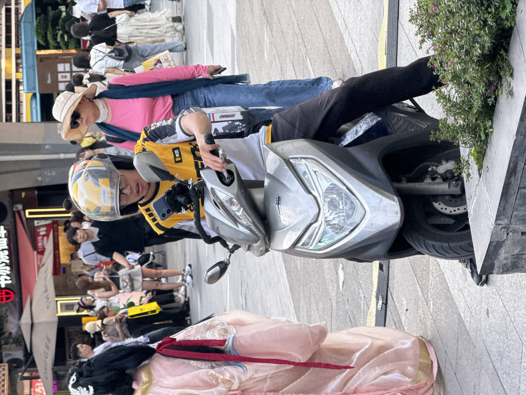 A pedestrian zone scene in Yuzhong District, China. A person in traditional East Asian attire is lying on the ground. A person on a parked scooter wearing a helmet, yellow vest, and black pants is visible. Pedestrians surround the scooter and the person lying on the ground. A person wearing a sunhat and pink shirt is partially visible behind the scooter. The sidewalk consists of concrete tiles. A bush in a concrete planter sits to the right. Buildings are visible in the background with Chinese characters on the storefronts.