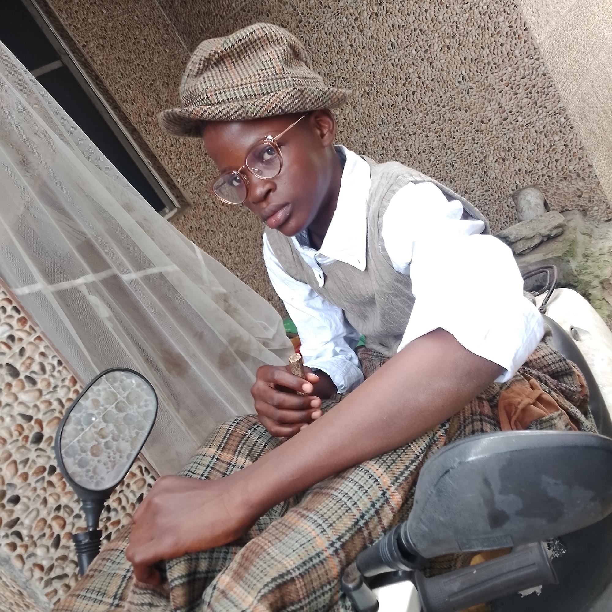 A young person, appearing to be male, is seated outdoors on what seems to be a motorcycle. They are wearing a light blue collared shirt, a grey knitted vest, and a plaid flat cap and trousers. Round, gold-rimmed glasses are perched on their nose. Their left hand is holding a small, light-brown cylindrical object, possibly a piece of wood. The background consists of a textured wall made of small, light brown pebbles and a white sheer curtain. To the left, a motorcycle mirror with a tiled reflective surface is visible. The overall setting suggests an urban environment, possibly in Ado, Nigeria, given the architectural style and materials used in the background. The lighting indicates it is daytime.