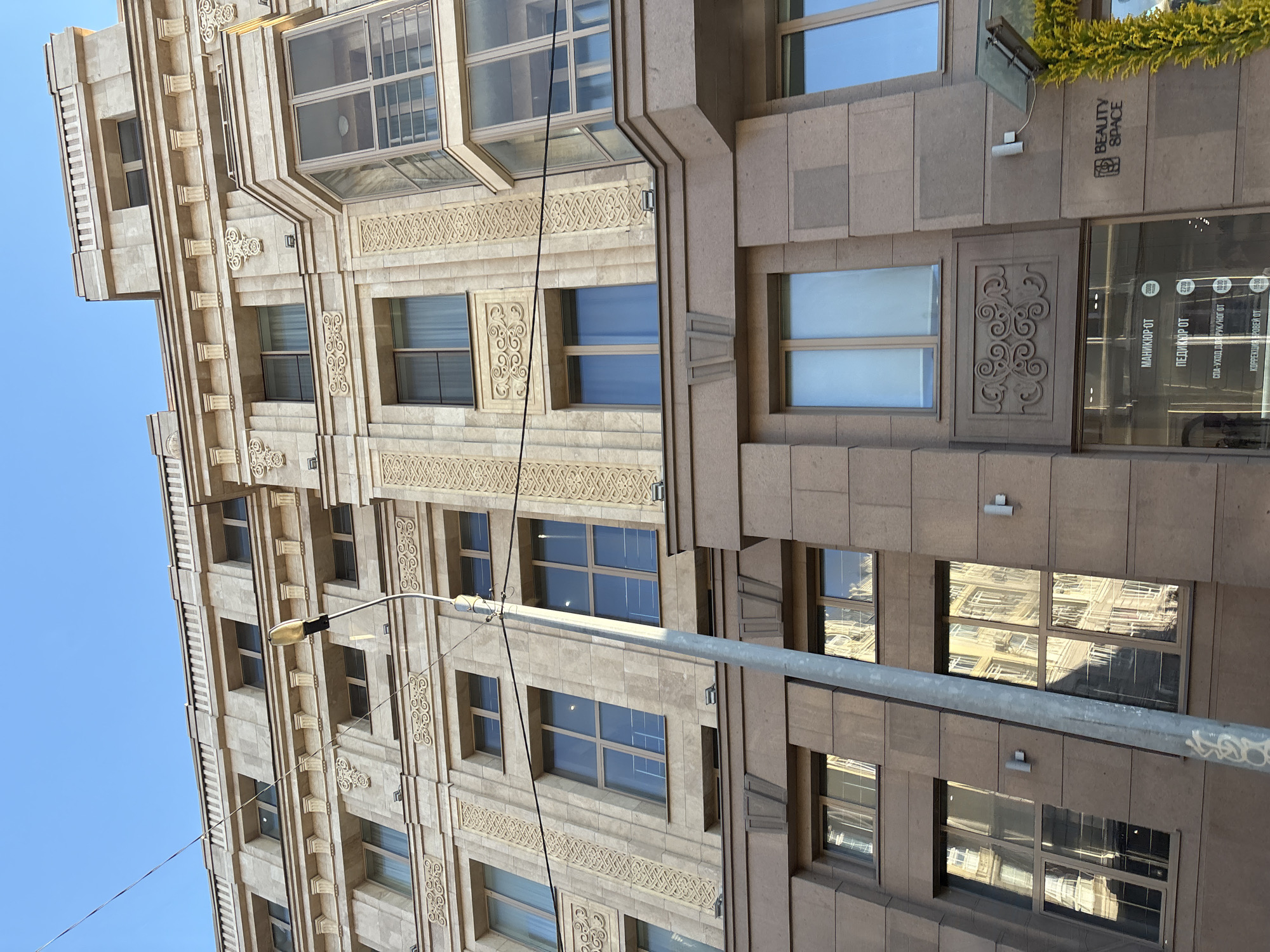 This photograph shows a portion of a building exterior, likely taken in Kazan, Russia, based on the Cyrillic signage. The building has a multi-story façade constructed from tan-colored stone or concrete. The architectural style features repeating window patterns interspersed with ornate decorative details, including carved floral or scroll designs. A lamppost runs vertically in the foreground, partially obscuring some of the building’s details. On the right side of the building, at ground level, there’s a business named "BEAUTY SPACE," with signage written in both English and Cyrillic, advertising manicure and pedicure services, as well as prices. Reflections of another building can be seen in the windows. A clear blue sky is visible on the left side. The photograph is taken at an oblique angle, looking upward at the building.