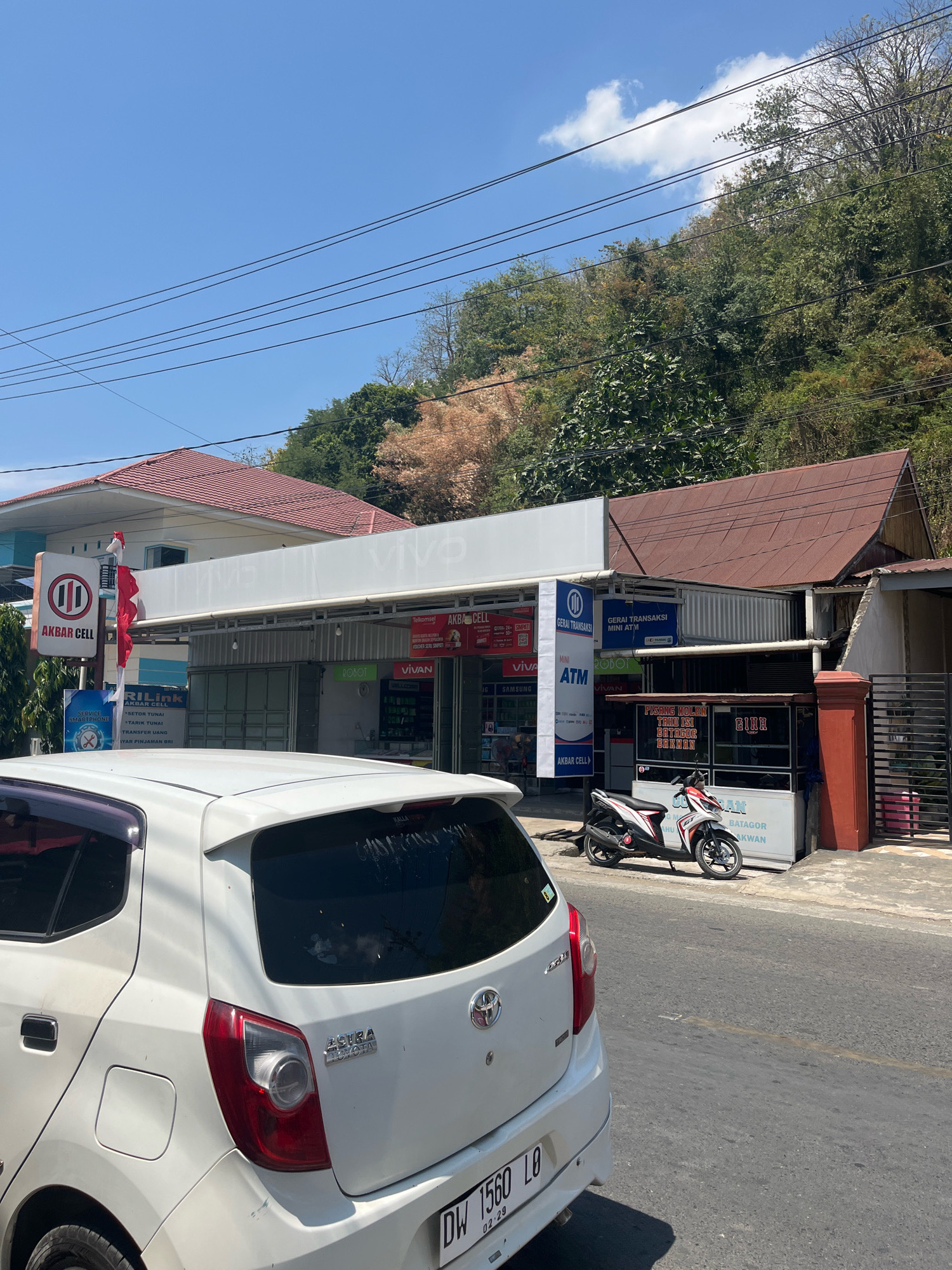 In Sengkang, Indonesia, a white car is parked on a street in front of a row of shops. The shops are a mix of businesses, including a cell phone store called "AKBAR CELL" and a convenience store. A motorcycle is parked in front of one of the shops. The shops are built along a street that appears to be on a slight incline, with a hill covered in trees and vegetation rising behind them. Power lines crisscross the sky above the shops and the street. The sky is clear and blue with a few scattered clouds. The car is a modern hatchback, and the motorcycle is a scooter-style vehicle. The shops have various signs advertising services and products. The overall scene depicts a typical urban streetscape in a tropical climate.