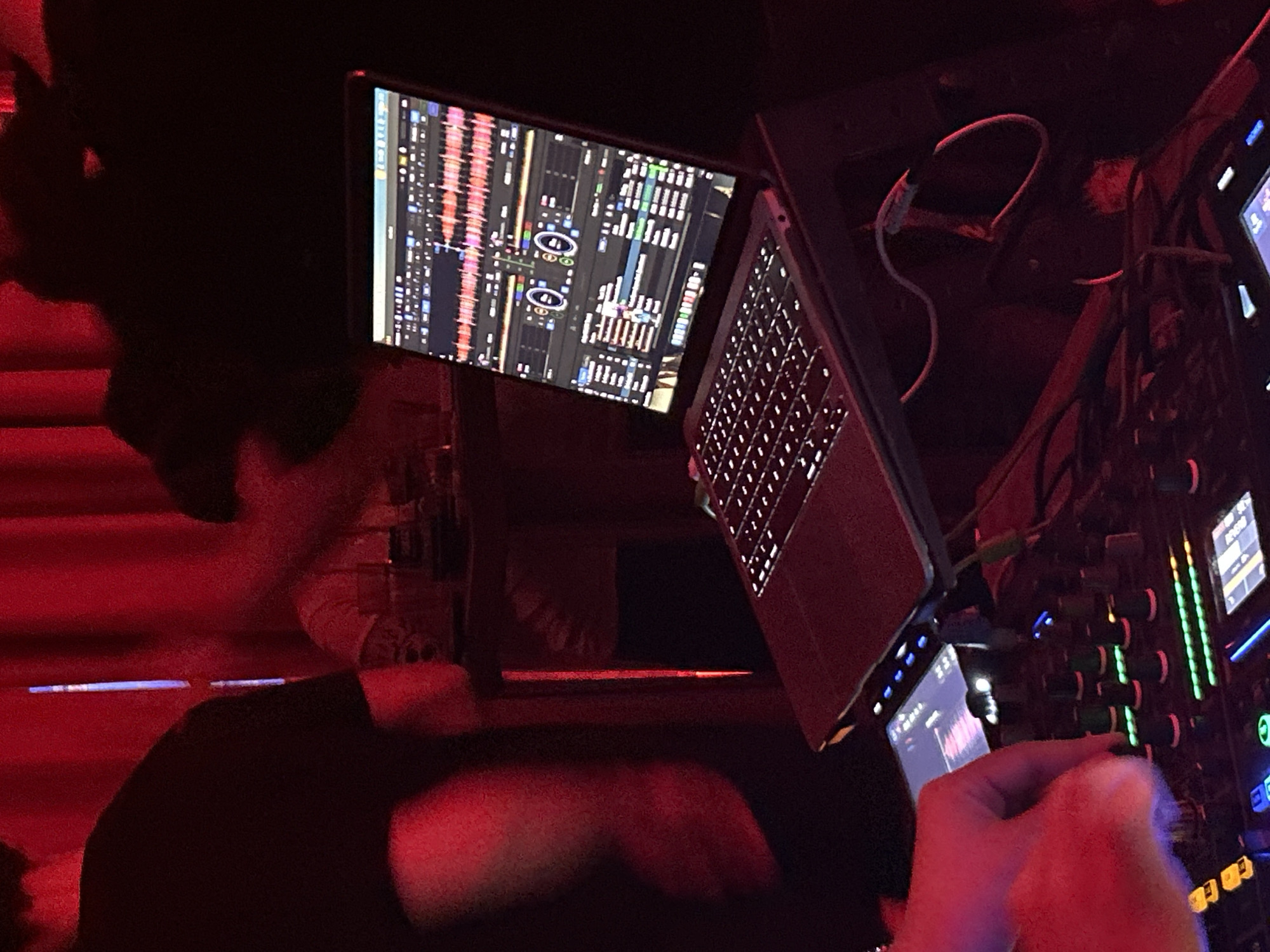 The image depicts a close-up view of a DJ setup in a dimly lit indoor venue, illuminated primarily by red ambient lighting. In the foreground, a human hand, distinctly highlighted by the red light, is actively manipulating a knob on a professional DJ mixer or controller. This device features an array of illuminated buttons and faders, exhibiting green, blue, and yellow lights. Above the controller, an open MacBook Pro laptop is positioned on a stand, its screen displaying a digital DJ software interface with prominent audio waveforms, track information, and virtual mixing controls. Connectivity cables are visible, linking the laptop to the mixer. Blurred silhouettes of at least two additional individuals are discernible in the background, with one person in a dark t-shirt partially visible on the left side of the frame. The background also reveals red curtains. This scene is situated in Varna, Bulgaria.