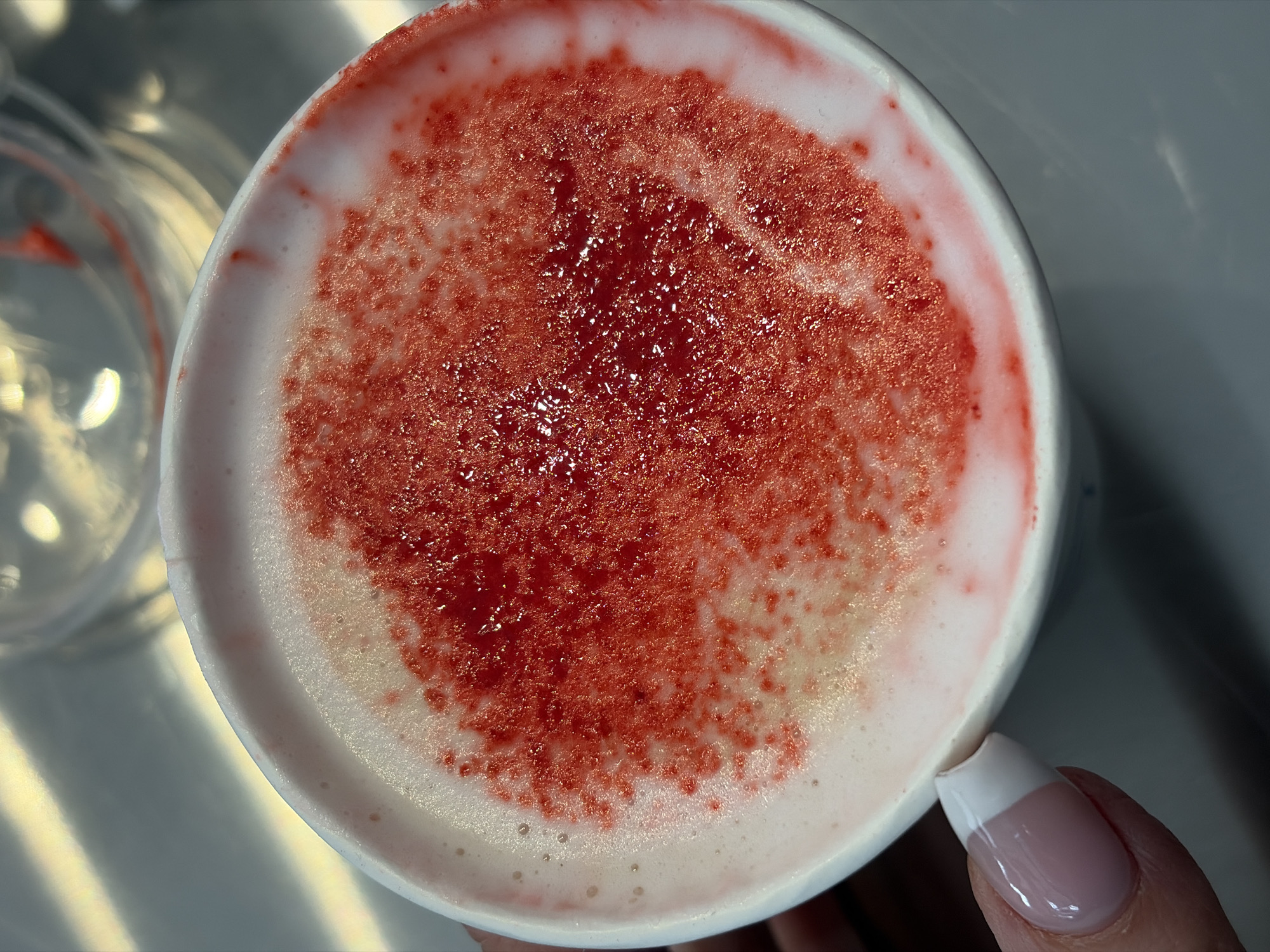 A top-down, close-up shot shows a full white coffee cup filled with a light pink froth base and topped with a layer of textured, deep red liquid. The surface of the red liquid appears slightly shiny and viscous, with some areas appearing more transparent. Light reflects from the liquid, creating highlights. The cup appears to be held by a hand, with a single finger visible. The fingernail is manicured with a white tip on a pink base. An out-of-focus glass or container can be seen in the background, slightly to the left of the cup. The surface on which the cup sits is a light gray color, possibly a table, with some light reflection creating variations in tone. The scene's location in Saint Petersburg, Russia, cannot be determined from the image.