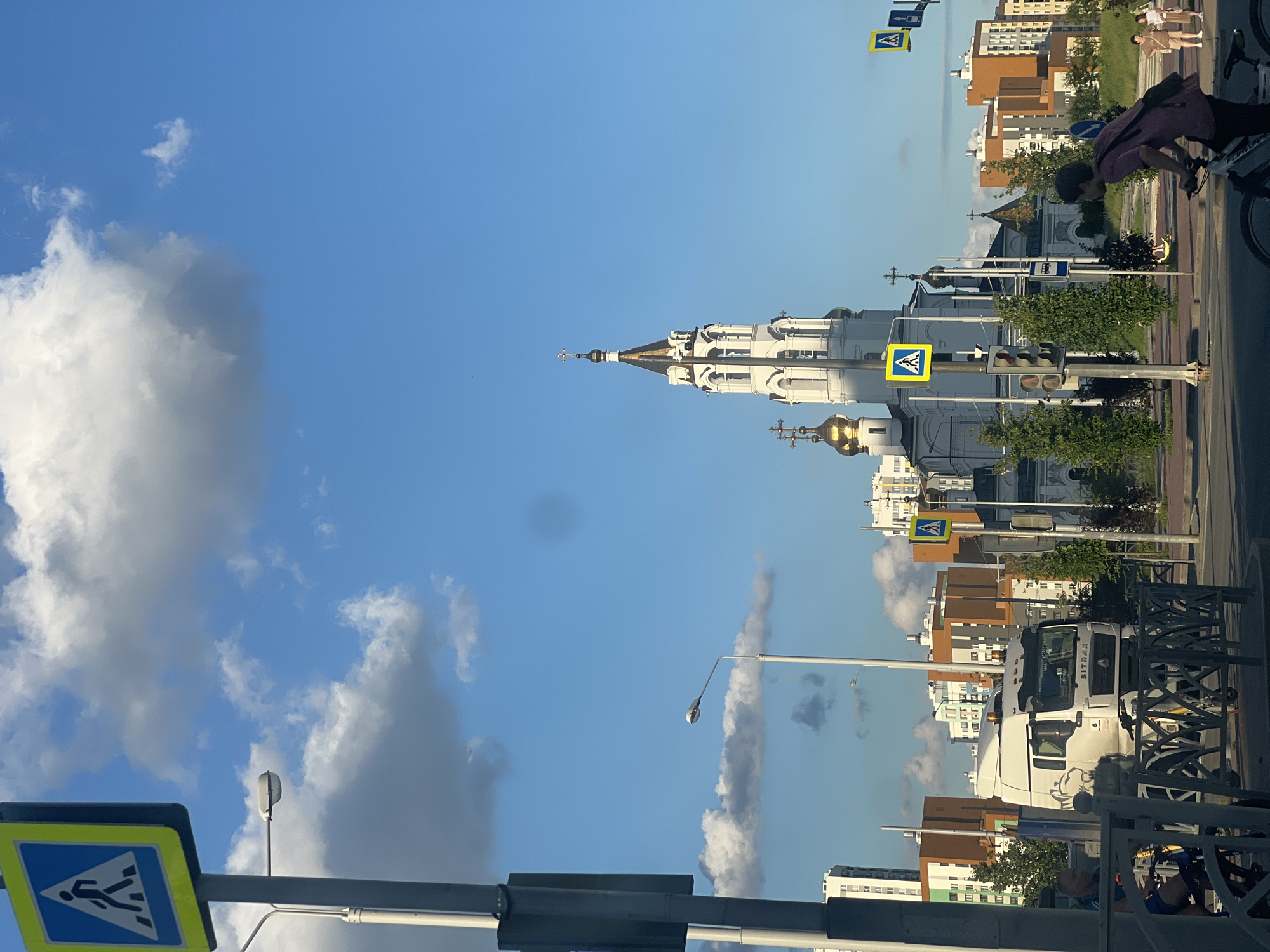 This is an eye-level, outdoor, daytime photograph taken in Yekaterinburg, Russia. In the foreground, a man is riding a bicycle from right to left across a street. Behind him, a white truck with "SITRAK" written on the front is parked. A white church with gold domes and a tall white steeple is visible in the background. The sky is blue with scattered white clouds. Buildings with orange and white facades are visible on the right side of the image. A pedestrian crossing sign is visible on the left side of the image, and another one is visible on the right side, closer to the church. The scene appears to be a street in a modern urban area, likely a residential or mixed-use district, given the presence of apartment buildings.