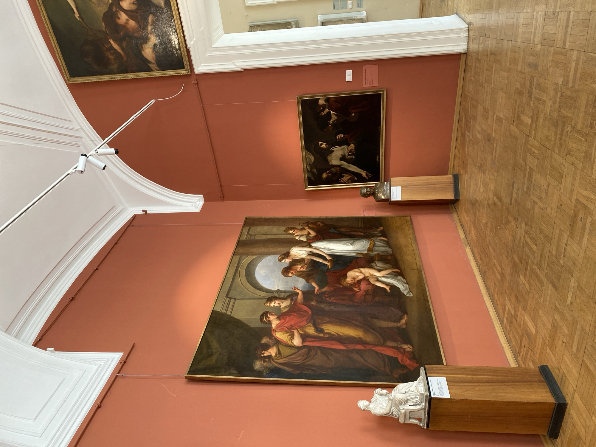 The image shows a museum interior in Unknown, Unknown. The walls are a reddish-orange color, and the floor is made of wood parquet. Three paintings hang on the wall. The largest painting depicts a crowd of people in classical attire, with figures in robes and a dramatic scene unfolding. A smaller painting shows a darker scene with figures in dark robes and a body being held. The third painting is partially visible and appears to depict a similar scene. In front of each of the two large paintings, there is a wooden pedestal topped with a white statue or bust. The environment suggests a formal art museum setting.