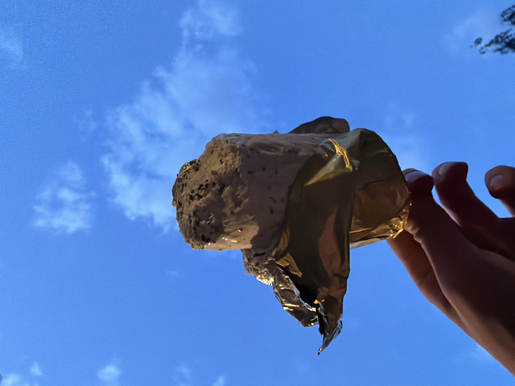 A hand holds an unwrapped ice cream sandwich against a bright blue, slightly cloudy sky. The ice cream sandwich is partially eaten, exposing the ice cream within. The wrapper is crumpled. The location is not identifiable as Moscow, Russia, without additional information.