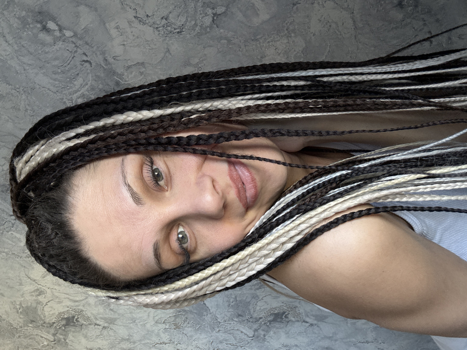 A close-up, rotated portrait features a woman with prominent multi-toned box braids. Her head is positioned to the left side of the frame, with her body extending to the right, suggesting she is lying down. Her hair consists of numerous thin braids in alternating shades of black, dark brown, and off-white/light blonde, framing her face and cascading across the upper part of her body.
The woman has fair skin, a gentle expression, and a slight, closed-mouth smile. Her light-colored eyes, possibly green or hazel, are open and directed towards the viewer. Her eyebrows are well-defined and dark. She appears to be wearing a light-colored, possibly white or light grey, top, with a strap visible on her right shoulder.
The background is a textured, mottled grey surface, resembling concrete or a painted wall, which provides a neutral backdrop that highlights her hair and face. There are no other objects or visible text in the image. The lighting is soft and even, illuminating her features without harsh shadows. The overall mood is calm and inviting.