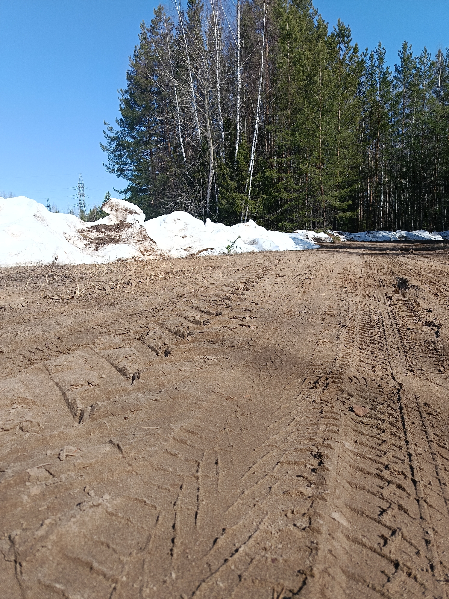 The image depicts a dirt road in Dubovaya, Russia. Tire tracks are visible in the foreground. Piles of snow line the left side of the road. In the background, a forest consisting of both evergreen and birch trees is present. Power lines are visible in the distance on the left. The sky is clear and blue. There are no people visible in the image.
