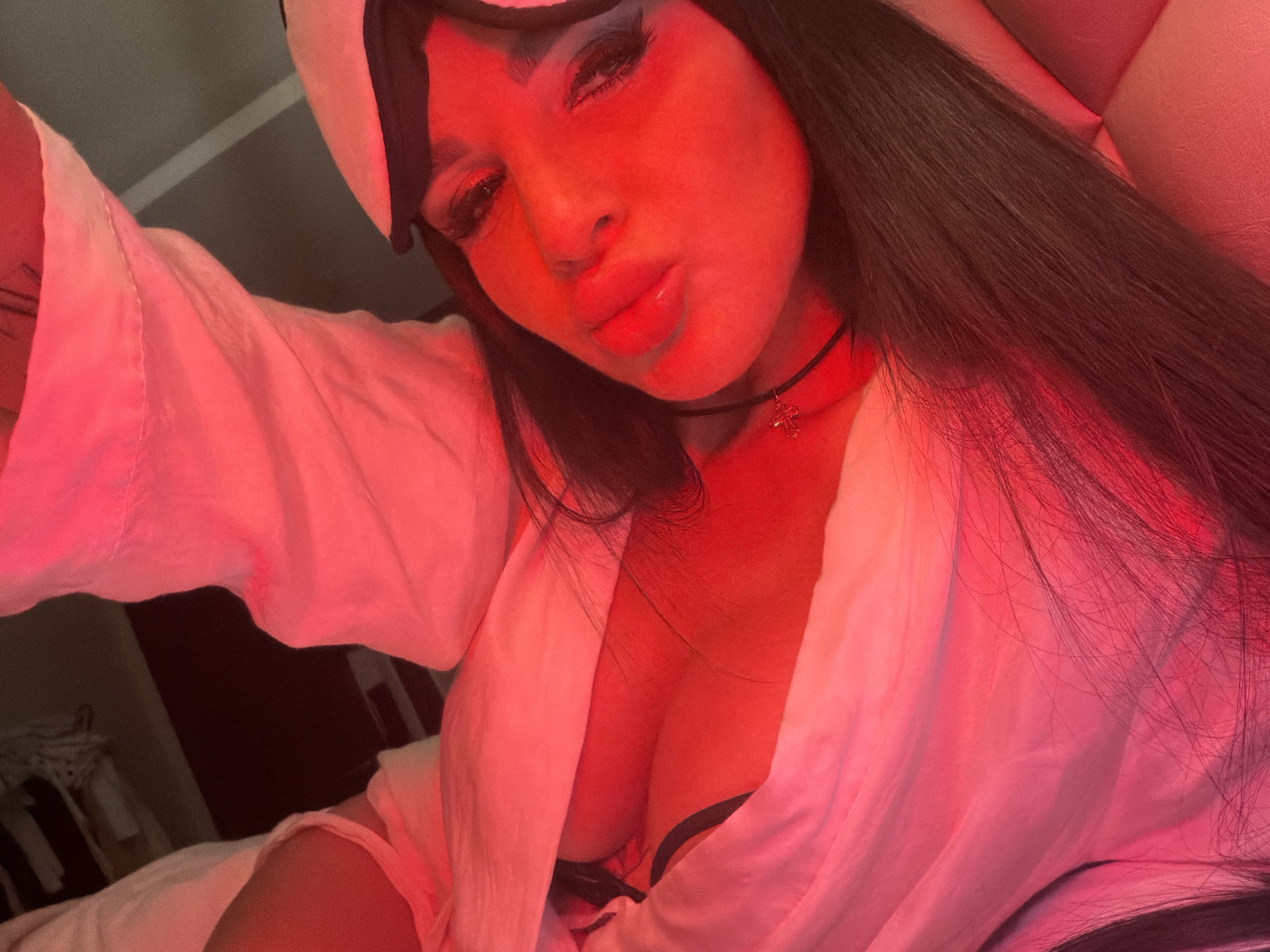 A woman with long, dark hair is posing in a low-angle selfie. She is wearing a light-colored robe and a sleep mask is pushed up on her forehead. She has heavy makeup on, with dark eyeshadow. She is wearing a choker necklace with a small pendant, possibly a cross. Her lips are pursed. The lighting is predominantly red, making the colors appear saturated. The background is out of focus, suggesting an indoor setting. The presence of a bed and a robe implies a bedroom environment. The location in Boreti, Montenegro, is not evident.