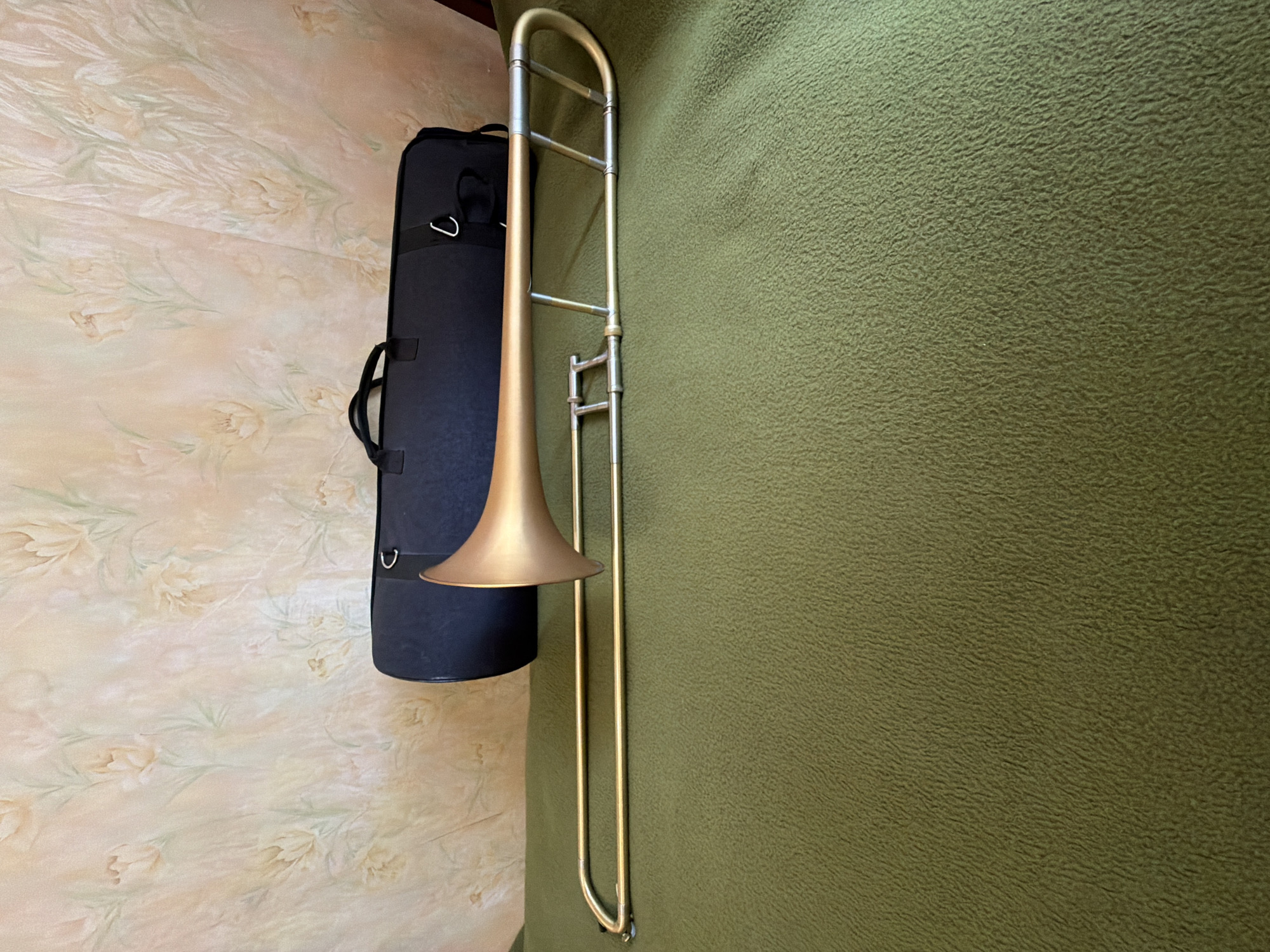 The image shows a golden brass trombone and its black soft carrying case laid out on two distinct textured surfaces. The trombone is positioned horizontally, with its bell facing towards the left and its slide extending across the frame. To the left of the trombone, the black case is visible, equipped with handles and a small hook.
The background is split between two different fabrics. On the left side, under the case and the trombone's bell, is a light-colored patterned fabric, possibly a bedspread, featuring subtle floral designs in light peach, pink, and green tones. The right side of the image, where most of the trombone rests, is covered by a textured, dark olive-green fabric, resembling fleece or a heavy blanket.
No activity is taking place; the objects are static, likely displayed indoors. The photograph is taken from an overhead perspective. There is no visible text in the image. While the provided location context is Kyiv, Ukraine, there are no specific visual cues within the image itself to confirm this.