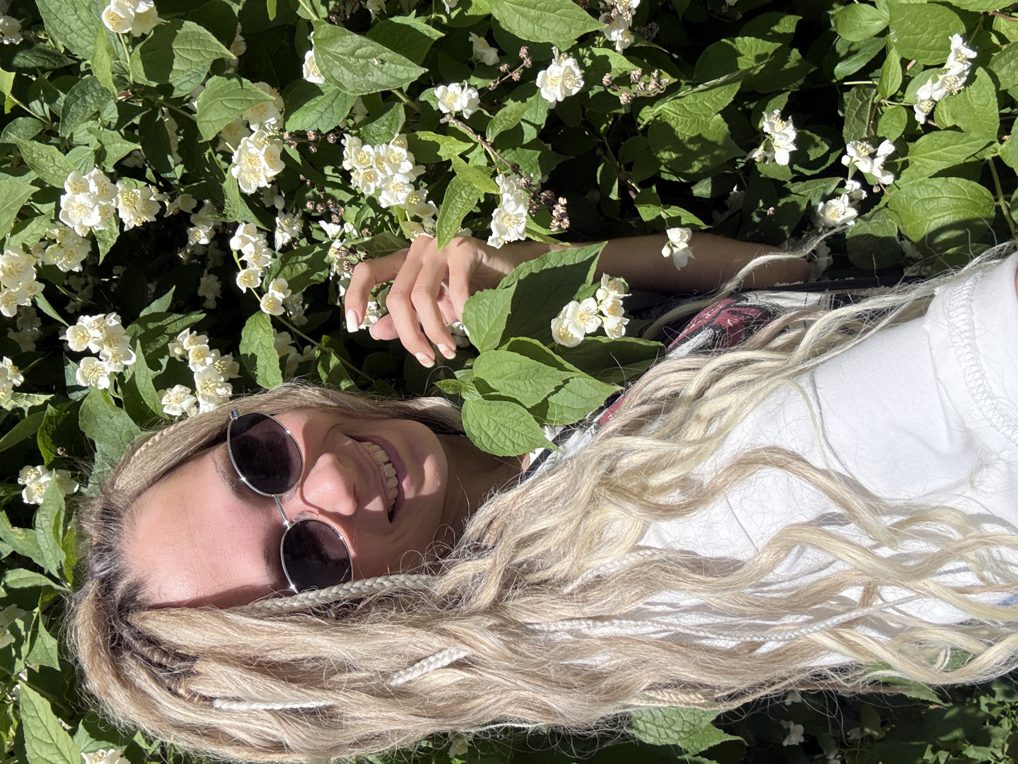 A young adult female is depicted outdoors in a sunlit environment. She stands with her body slightly angled, looking towards the camera with a wide smile. Her blonde hair is styled with a combination of loose waves, dreadlocks, and braids, some of which are lighter in color, almost white. She wears dark, oval-shaped sunglasses with silver frames. Her right hand, featuring manicured nails with light polish, is gently placed among the green leaves of a dense flowering bush that forms the background. The bush is covered in numerous small, white flowers, each with multiple petals and visible stamens, consistent with species like jasmine or mock orange (*Philadelphus*). The specific location in Rusanovo, Russia, cannot be visually confirmed from the image data alone.
