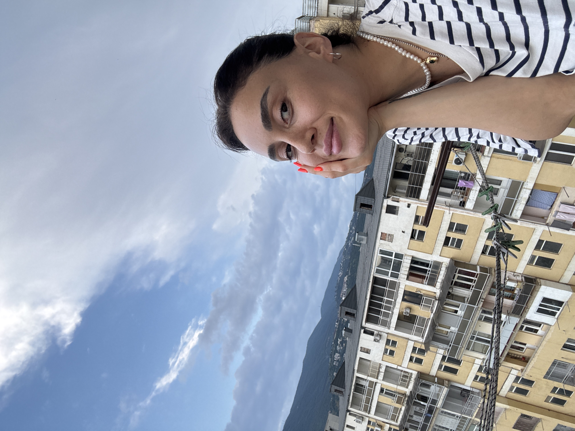 In this close-up, outdoor, eye-level shot, a young woman with dark hair and eyebrows is pictured from the shoulders up, leaning her head on her right hand. She is wearing a striped t-shirt and a pearl necklace with a gold pendant. Her nails are painted a bright orange. She is looking towards the camera with a slight smile.
In the background, there is a multi-story yellow building with balconies, windows, and air conditioning units, characteristic of Soviet-era architecture found in Tbilisi, Georgia. The building is situated on a hillside, with other buildings and a mountain range visible in the distance under a partly cloudy blue sky. Laundry is visible hanging on clotheslines attached to the building, with green clothespins securing the items. The overall scene suggests a residential area in Tbilisi.