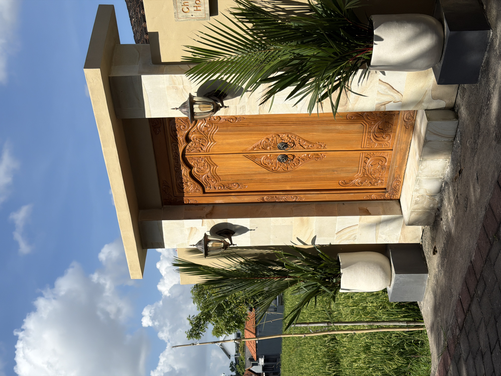 The image captures the entrance of a building, likely a villa or guesthouse, in North Kuta, Indonesia. A pair of intricately carved wooden doors are flanked by light beige stone columns and topped with a matching lintel. On either side of the door are potted plants with palm-like foliage. Two ornate metal lanterns are mounted on the wall above the doors. The sky is visible in the background, partially obscured by the building. The outdoor setting includes greenery, possibly a rice paddy field, suggesting a rural or semi-rural location. The overall composition is focused on the building's entrance and its architectural details.