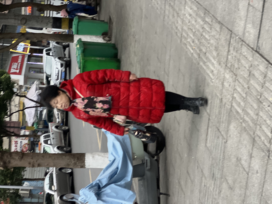 This image depicts an urban street scene in Jiangjin, China. An older adult East Asian female is walking from right to left on a gray paved sidewalk composed of rectangular and square tiles, including sections of tactile paving. She is dressed in a long, red, quilted winter coat, black pants, and black boots. A dark scarf with a pink and white floral pattern is worn around her neck. Her short hair is dark and curly. She holds a small, light-colored purse with a chain strap in her right hand, along with a darker, patterned bag. To her left, a scooter or small motorcycle is partially covered by a light blue tarp, with a patterned bag hanging from its handlebars. Further left, a tree trunk with a white-painted base is visible. To her right, three green public waste bins are present, two situated side-by-side. In the background, an asphalt road displays multiple vehicles, including a silver sedan and several silver pickup trucks. Buildings with commercial facades, featuring a red sign with white lettering and another with blue neon lighting, line the far side of the street. The lighting suggests an overcast daytime environment.