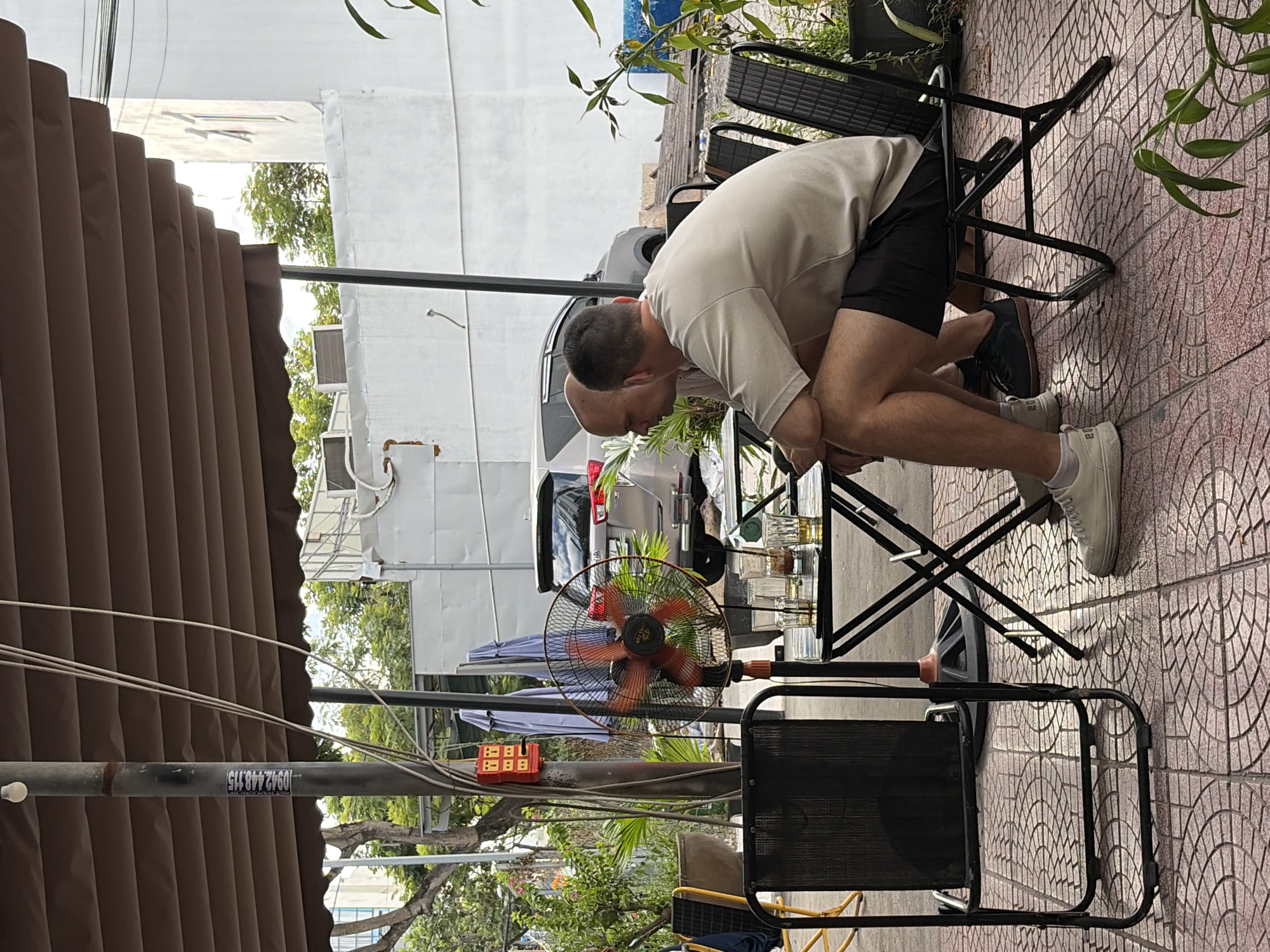 Two men are seated at a small table under a brown awning at an outdoor cafe in Da Nang, Vietnam. The man on the left, with a shaved head, is looking down at his phone. The man on the right, with dark hair, is wearing a light-colored t-shirt and black shorts and appears to be looking at something on the table. On the table are several glasses, some with beverages. A black metal chair with a mesh seat is in the foreground to the left, and another similar chair is to the right, occupied by the second man. A floor fan with an orange frame is positioned behind the table. The cafe appears to be situated on a tiled patio, with potted plants and a silver car visible in the background. The overall lighting suggests it is daytime.