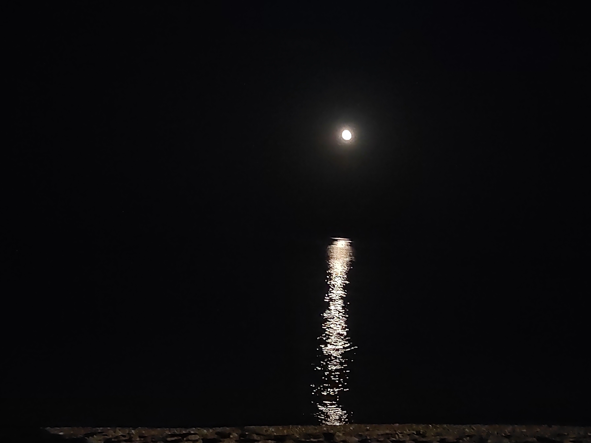 A full moon shines brightly in a dark night sky over the Adriatic Sea in Porto Recanati, Italy. Its reflection creates a shimmering, elongated path of light on the water's surface. In the foreground, a rocky shore is visible, hinting at the edge of the land. The scene is serene and undisturbed, with no visible people or activity. The atmosphere is one of natural beauty and tranquility.