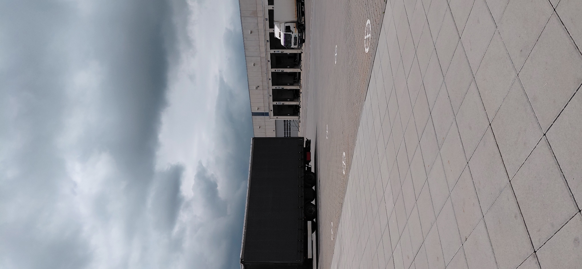 The image depicts an outdoor scene at a logistics facility. In the foreground, a paved area with large, square concrete tiles leads towards the mid-ground. Scattered across this area are circular white markings, likely indicating parking or loading zones.
Dominating the left side of the mid-ground is the rear of a dark-colored, large truck trailer, a semitrailer, parked lengthwise. Its wheels and lower chassis are visible.
In the background, a long, modern warehouse building stretches across the frame. It features a series of loading bays, each marked with a number (e.g., 69, 68, 67, 66, 65, 64, 63). Several of these bays appear to be closed with roll-up doors, while others are open. A white truck cab, possibly attached to a trailer not fully visible, is parked at one of the loading bays on the right. The warehouse is constructed with a light-colored, possibly concrete, facade and has a blue vertical accent strip above the numbered bays.
The sky above is overcast with heavy, dark grey clouds, suggesting an impending rain or a generally gloomy weather condition.
The scene is devoid of any visible people. The overall impression is one of a functional industrial or logistics hub under a cloudy sky. This scene is located in Bozhurishte, Bulgaria, at a logistics complex.