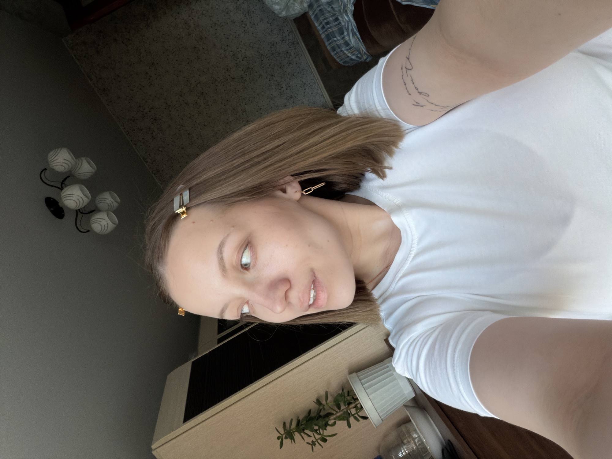 A young woman with light brown hair is angled in the frame, wearing a white T-shirt. She has a gold clip in her hair and gold earrings. She is indoors. In the background there is a light colored cabinet and a potted plant. A set of modern wall lamps can be seen in the background. The location is Razdolie, Russia.