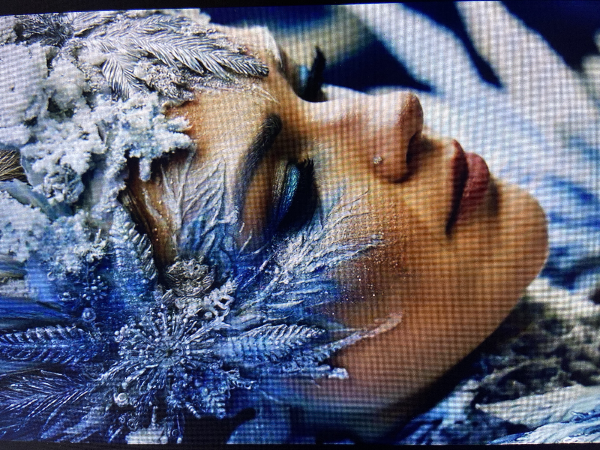 The image is a close-up of a woman's face adorned with elaborate, frosty, blue and white makeup and costume elements, resembling ice and snowflakes. Her eye makeup is dark blue and shimmery, with long eyelashes. She has a small nose stud. The overall aesthetic is that of an ice queen or winter spirit. There is no discernible location in Stavropol, Russia, or any other specific city, as the image is highly stylized and does not contain any recognizable landmarks or contextual environmental clues.