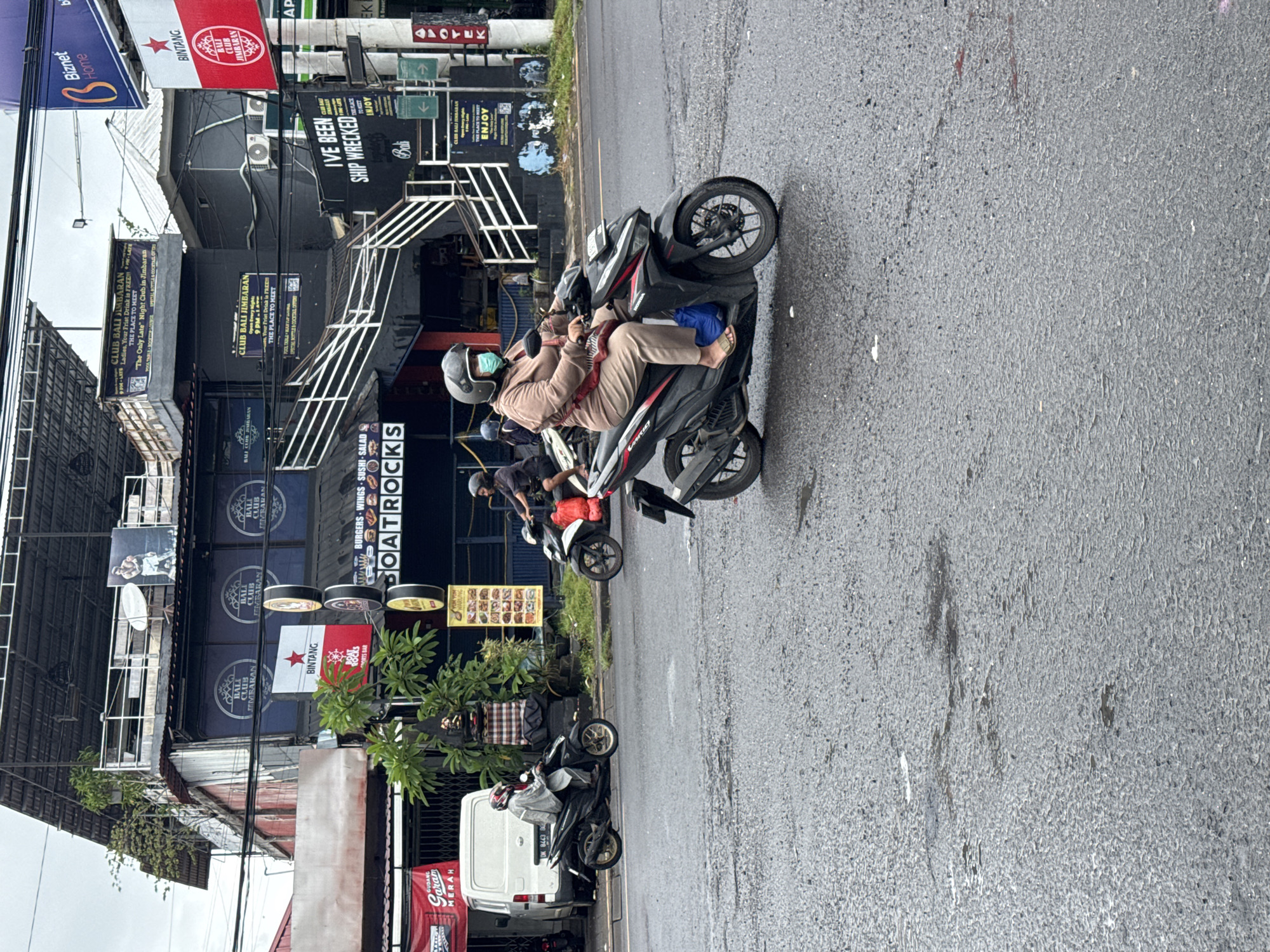 This image captures a street scene in Kuta Selatan, Indonesia, characterized by an overcast sky and a wet asphalt road, suggesting recent rainfall. Several individuals are seen on scooters and motorcycles. In the foreground, a person wearing a helmet, a face mask, a light brown top, and beige pants sits on a black Honda scooter, with a blue object tucked into the footrest area. Other scooter riders are visible further down the road, with one wearing a black shirt and another with a white cover-up.
The street is flanked by multi-story commercial buildings. Numerous signs advertise various establishments. Prominently featured are "CLUB BALI JIMBARAN," billed as "The Only Late Night Club in Jimbaran," offering "Ladies, Your First Drink is FREE!!!" and open from "9 PM - LATE." Another sign reads "BOATROCKS" advertising "BURGERS - WINGS - SUSHI - SALAD" and a "SPORTS BAR." Other visible texts include "BINTANG" (a beer brand), "Biznet Home," "IVE BEEN SHIP WRECKED," "Bali," "ENJOY," "APOTEK" (pharmacy), "WARUNG," and "GUDANG GARAM MERAH" (a cigarette brand). A white van with the license plate "DK 8443" is parked by the roadside. The scene depicts a lively urban environment with diverse commercial offerings.