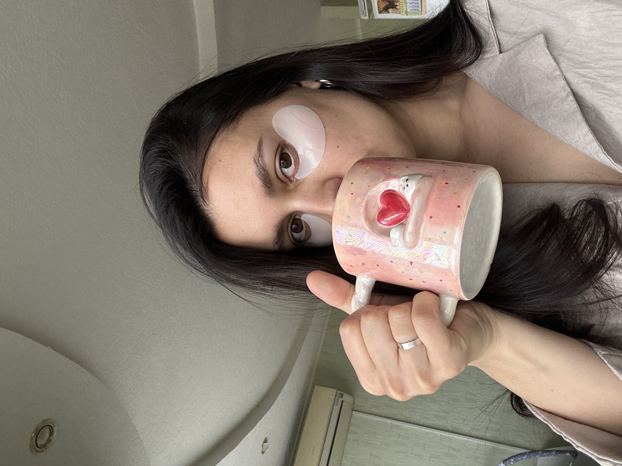 A young woman with dark hair and brown eyes is holding a pink mug with a bear and a red heart on it. She is wearing an eye mask under her left eye. A ring is visible on her left hand. The background is a light-colored wall, with an air conditioning unit visible. The location is Chaikovsky, Russia.