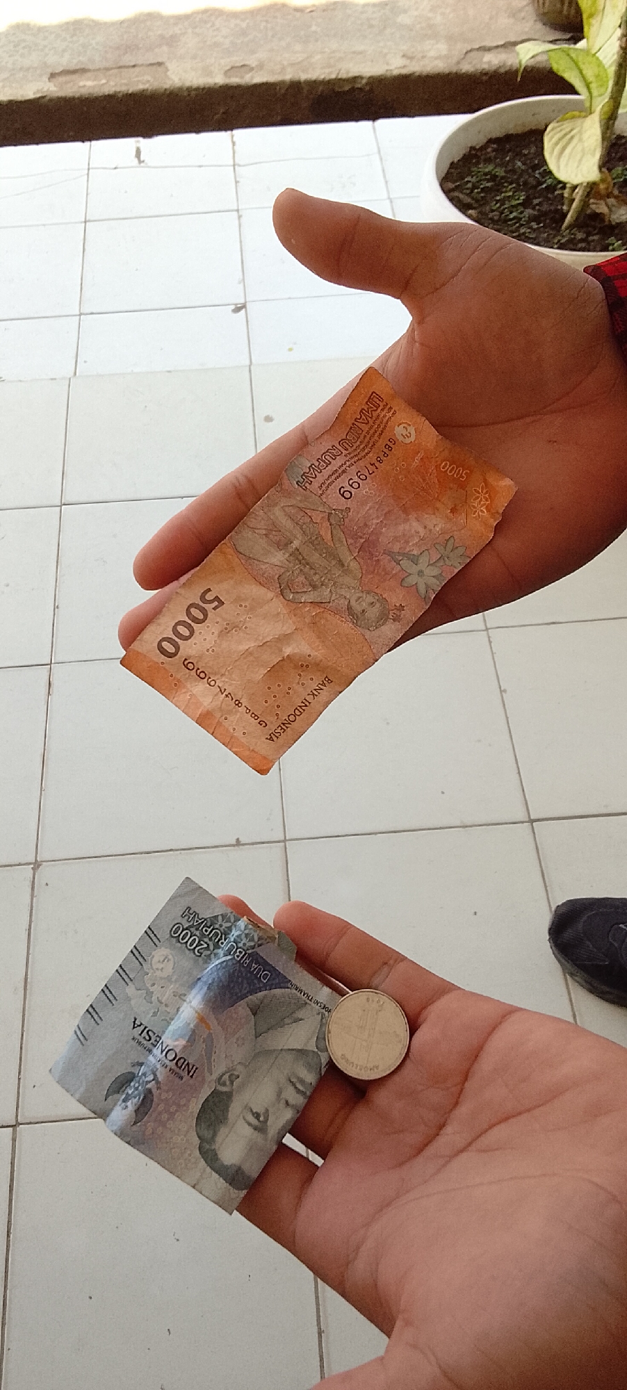 In Jepara, Indonesia, a person's hands are holding two Indonesian Rupiah banknotes and one coin. The top banknote is orange and appears to be a 5,000 Rupiah note, with the number "5000" prominently displayed and the text "BANK INDONESIA" visible. The lower banknote is blue and shows "2000" and "INDONESIA" and "DUA RIBU RUPIAH." A silver coin, possibly of a smaller denomination, is placed on the lower banknote. The hands are positioned over a tiled floor. In the background, a potted plant with green leaves is visible. The scene appears to be outdoors.