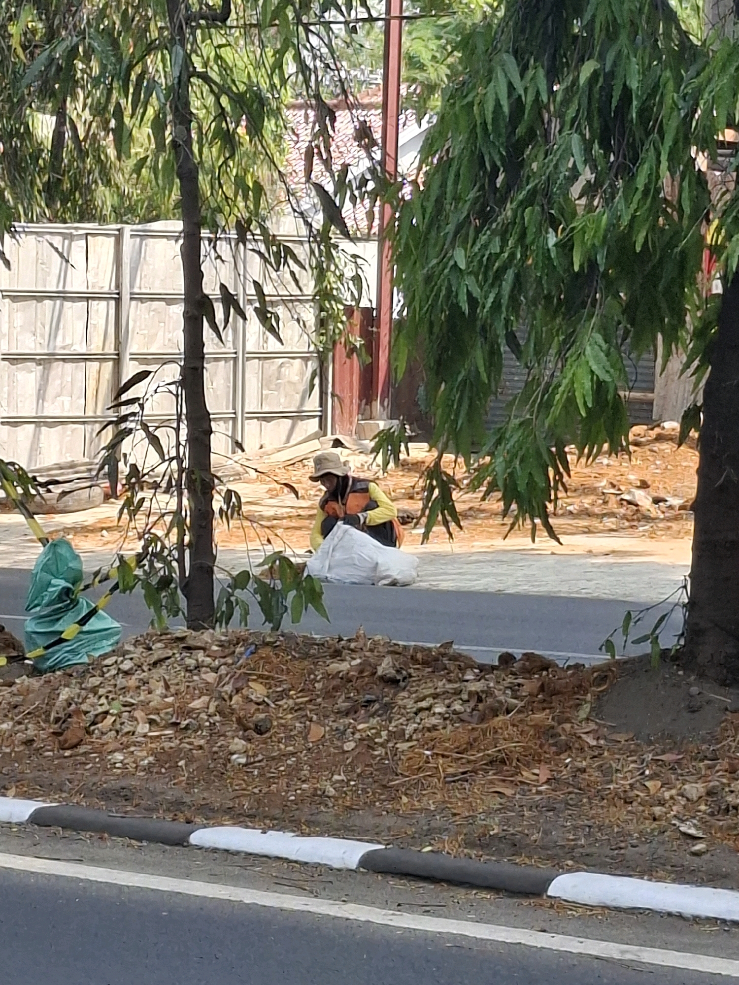 In Sukabumi, Indonesia, a person wearing a reflective vest and a hat kneels on the side of a road, working with a large white bag. Behind them, a temporary fence constructed of metal sheeting encloses a construction area, with exposed dirt and scattered debris. Several trees with dense green foliage are present in the foreground and background. A bright green tarp is visible on the left side of the image, secured with yellow and black striped tape. The road in the foreground has a white and grey painted curb. The scene appears to be an outdoor urban or suburban environment under daylight.