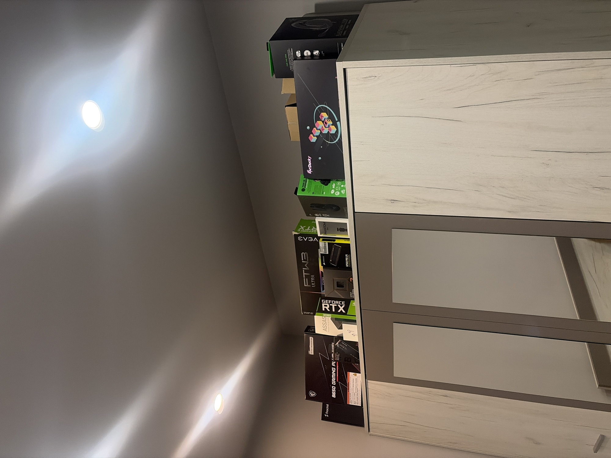 In Podgumer, Bulgaria, on the top of a modern wardrobe with light wood-effect doors, a collection of computer hardware boxes is arranged. The boxes, mostly black and green, are stacked from left to right, displaying prominent branding like "Fractal," "MSI," "EVGA," "GEFORCE RTX," and "Ducky." Among the boxes are also smaller items like a pair of white wireless earbuds. The overall scene is indoors, under a ceiling with recessed lighting that casts a glow across the room. The wardrobe's mirrored doors reflect the surrounding environment, adding a sense of depth to the image.
