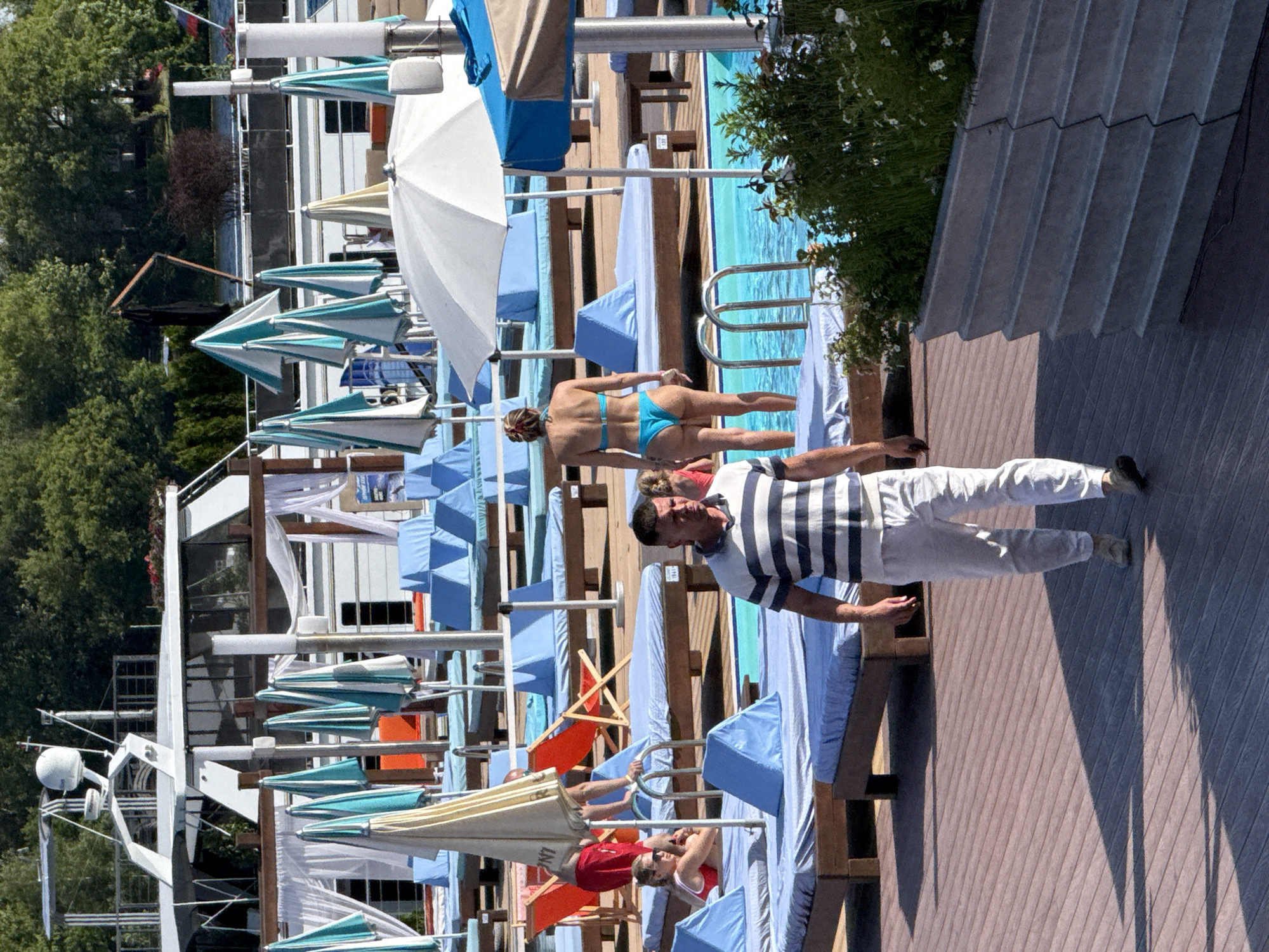 The image depicts a sunny day at an outdoor pool area, likely a resort or a hotel, with a focus on people relaxing. In the foreground, a man in a striped shirt and white pants walks across a wooden deck, his hand extended as if gesturing or reaching for something. Behind him, a woman in a blue bikini is positioned near the pool, seemingly enjoying the sun. The background is filled with rows of lounge chairs and umbrellas, many of them blue and white, suggesting a well-equipped leisure facility. A portion of the pool itself is visible, with clear blue water. Lush green trees and foliage are seen on the left side of the image, adding to the outdoor atmosphere. The scene is bathed in bright sunlight, casting distinct shadows on the wooden deck, indicating it is midday. The overall mood is relaxed and summery. There is no visible text in the image.
