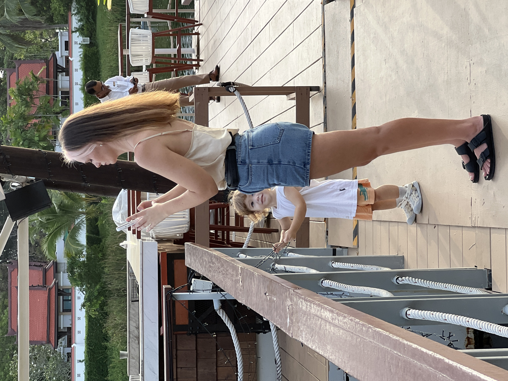 A young woman with long, ombre-colored hair is standing on a wooden deck, holding a smartphone in her hands and looking at it. She is wearing a light-colored satin camisole, a black belt, and a denim mini-skirt. A young child in a white t-shirt and orange shorts is standing to her left, looking up and to the right. Behind them, an Indian man in a white shirt and tan pants is looking at his phone, and in the background, there are white chairs and tables. The setting appears to be a waterfront area with a body of water and lush greenery.
