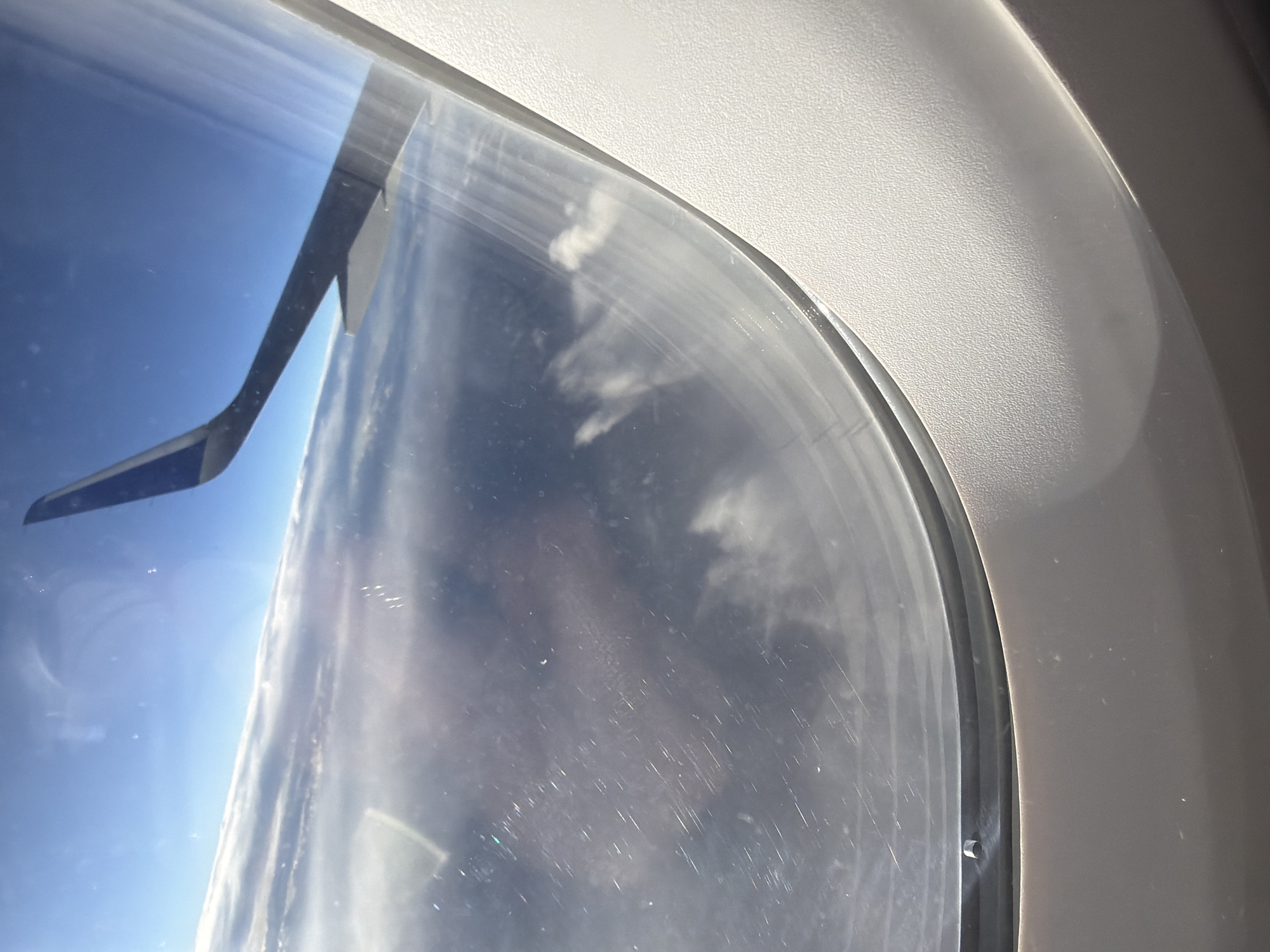 The image is taken from an airplane window, looking out at the sky and clouds. An airplane wing is visible on the right side of the frame, pointing towards the top right corner. The view outside the window consists of a vast expanse of clouds, with a clear blue sky above. The clouds are layered and appear to stretch as far as the eye can see. The airplane window itself shows some smudges and streaks, suggesting it may be dirty. The interior of the airplane cabin, including the window frame and surrounding paneling, is visible in the foreground and on the right side of the image. There are no people or specific landmarks visible that would allow for pinpointing a location within Denver, Colorado.