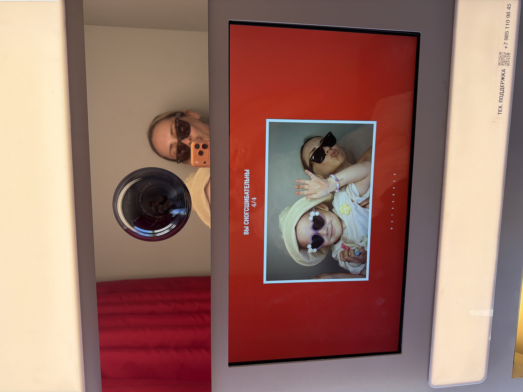 The image captures a selfie taken in a photobooth located in Khimki, Russia, showing both the reflection of the photographer and the photobooth's display screen.
In the upper portion, a woman is reflected, wearing a light-colored top, dark sunglasses, and a headband on her blonde hair. She holds an orange smartphone to her mouth, puckering her lips. The reflection reveals she is holding a Canon DSLR camera, with "Canon ZOOM LENS EF-S 18-55mm" visible on the lens. A red curtain is reflected on the left, and a light-colored wall on the right.
The lower portion displays the photobooth's screen, featuring a vibrant red background. White Russian text at the top reads "ВЫ СНОГСШИБАТЕЛЬНЫ" (You are stunning/awesome) and "4/4," indicating the fourth of four pictures. A framed photograph is centered on the screen, depicting a small child and an adult woman. The child, positioned on the left, wears a light-colored brimmed hat, a light-colored shirt, and heart-shaped sunglasses decorated with small white flowers, with her left hand raised. The adult woman, on the right, wears dark sunglasses and also puckers her lips. Her right arm, adorned with a colorful beaded bracelet, is visible around the child. White Russian text at the bottom of the screen reads "Ф О Т О К И О С К" (Photo Kiosk).
Below the screen, a white panel features a "ТЕХ. ПОДДЕРЖКА" (Tech Support) sticker, a QR code, and a Russian phone number: "+7 985 110 98 45."