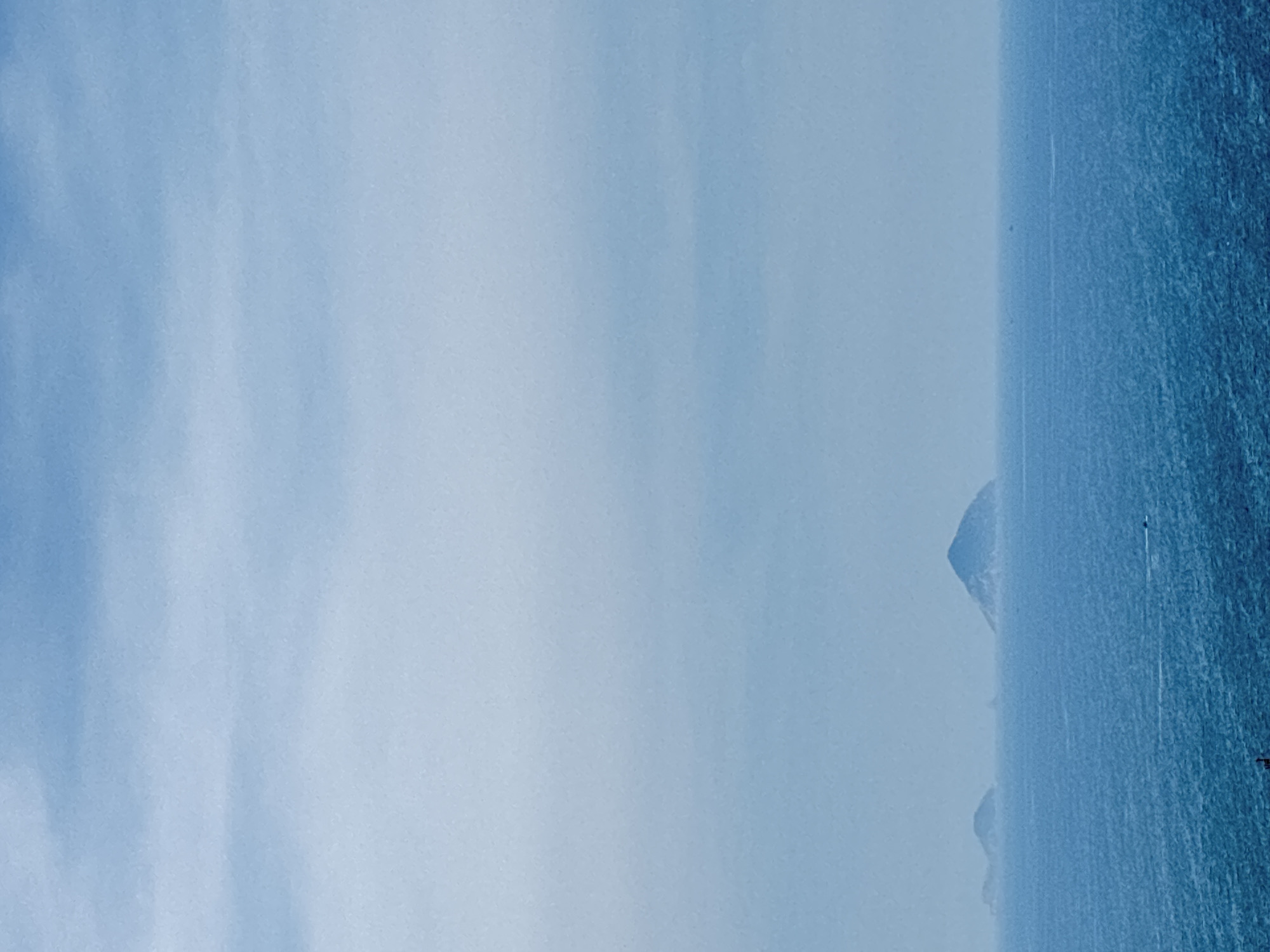 An aerial, full shot shows a vast expanse of blue ocean under a hazy, pale blue sky. The horizon is lined with several distant, low-lying islands. The largest island, in the center of the frame, is conical in shape and appears to be covered in vegetation. To the left of the largest island, two smaller islands are visible, also appearing to be covered in vegetation. A small, dark boat is visible in the bottom left corner of the frame. The sea is choppy, with visible waves. The overall impression is one of a remote and peaceful seascape.