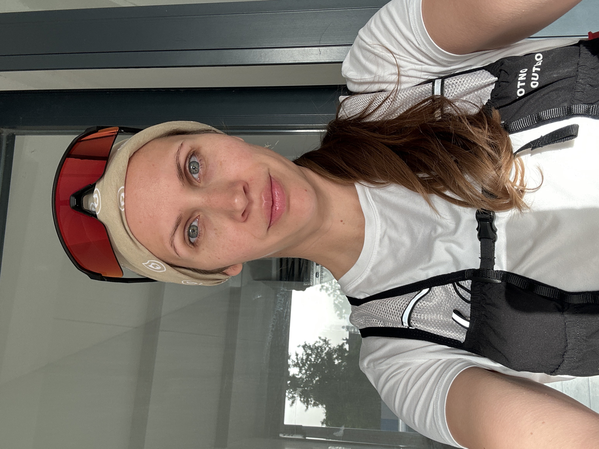 A woman with brown hair and blue eyes is posing for a selfie. She is wearing a white t-shirt, a hydration pack, and a beige headband with sunglasses pushed up on top. The background shows a window with a reflection of a tree and cloudy sky. The location is potentially Fethiye, Turkey, based on context. The subject is the woman. The objects include the t-shirt, hydration pack, headband, sunglasses, window, tree, and cloudy sky. No actions or interactions besides the posing are explicitly visible. The environment appears to be a building with a window looking outside.