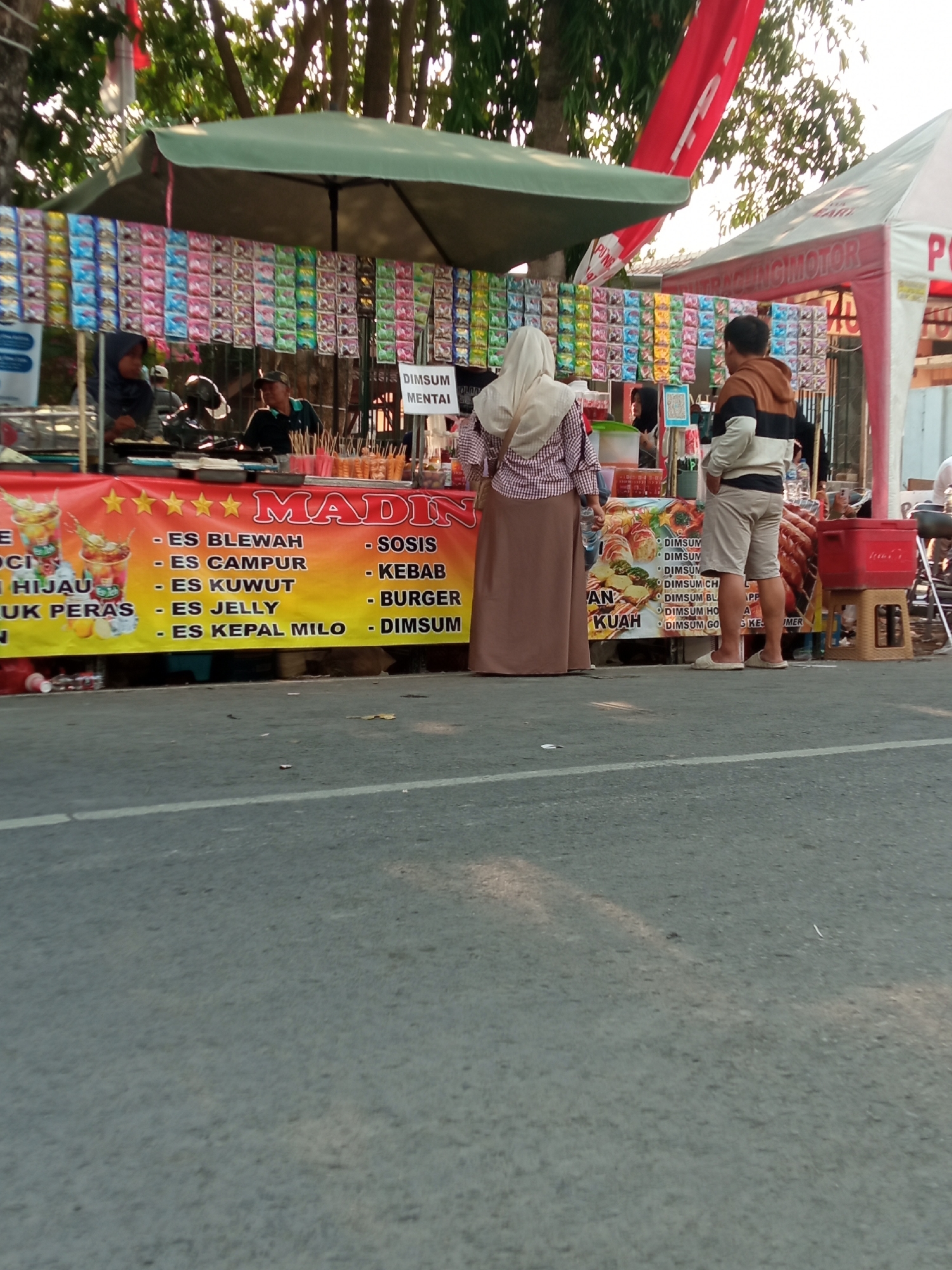 The image depicts a street food stall in Balun, Indonesia, on a sunny day. The stall is set up on the side of a road, with a colorful banner advertising "MADIN" and various food items such as "ES BLEWAH," "ES CAMPUR," "ES KUWUT," "ES JELLY," "ES KEPAL MILO," "SOSIS," "KEBAB," "BURGER," and "DIMSUN." Several people are visible at the stall. A woman wearing a hijab and a checkered shirt is interacting with a vendor, who is partially obscured by the stall. A man in a striped sweater and shorts is standing to the right of the stall, looking away from the camera. The stall is covered by a green awning, and a red tent is visible in the background. Rows of packaged snacks are displayed above the food banner. The ground is paved with asphalt, and a white line marks the edge of the road. Trees and foliage are present in the background, suggesting an outdoor environment. The overall scene captures a typical street-side food vendor operation in an Indonesian city.