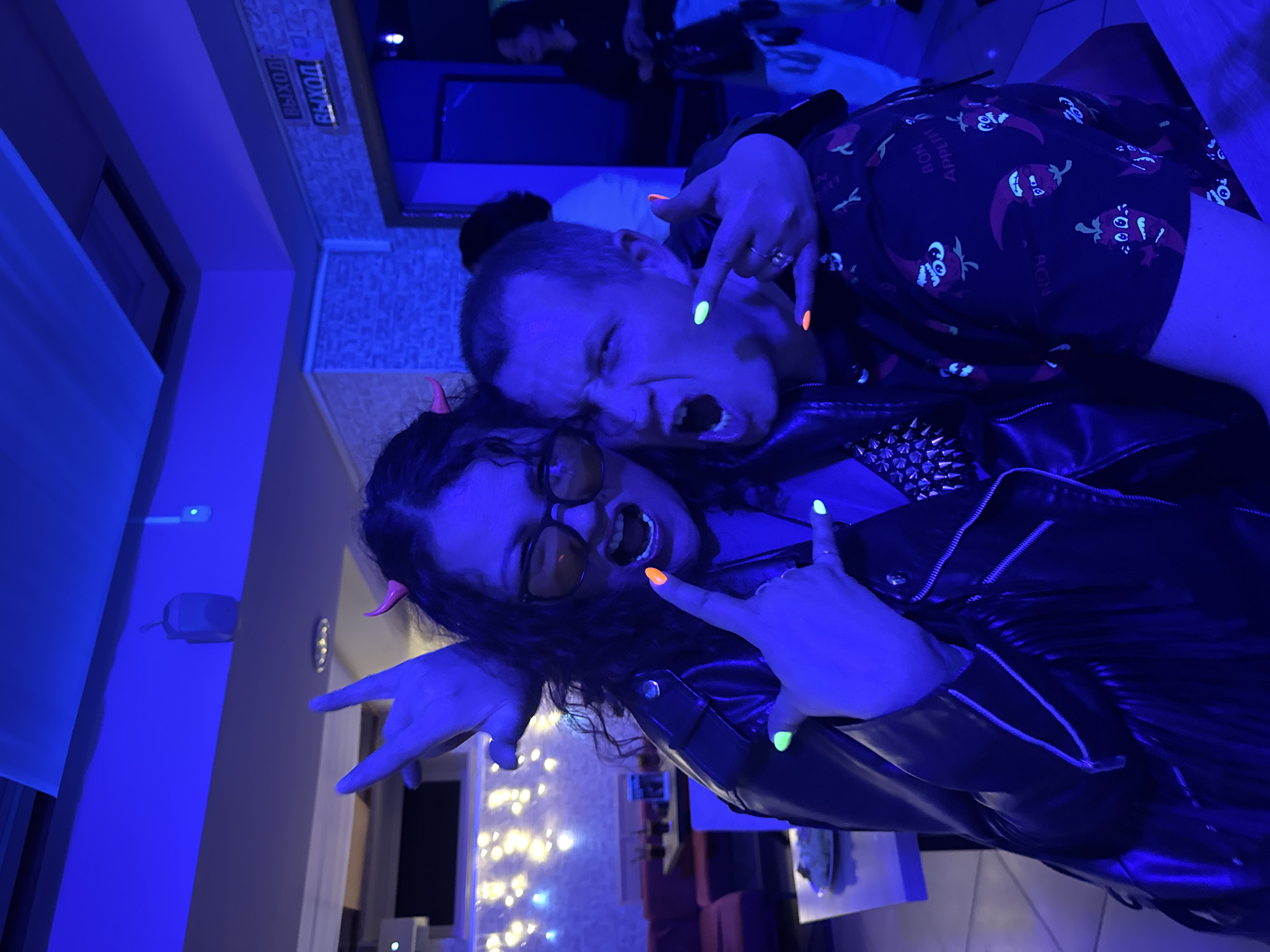 Two individuals are posing for a photograph under a blue light, possibly in a nightclub or entertainment venue. The man on the right has short hair and is wearing a black shirt with red chili pepper designs. He is making a face and hand gesture with bright green and orange painted nails. The woman on the left has curly dark hair and orange tinted glasses, with red devil horns. She is wearing a black leather jacket and also making a hand gesture with bright orange and green nails. The background includes a bar or serving area with illuminated bottles and a sign in Russian that may indicate an exit. The location is possibly in the city of Chaikovsky, Russia.