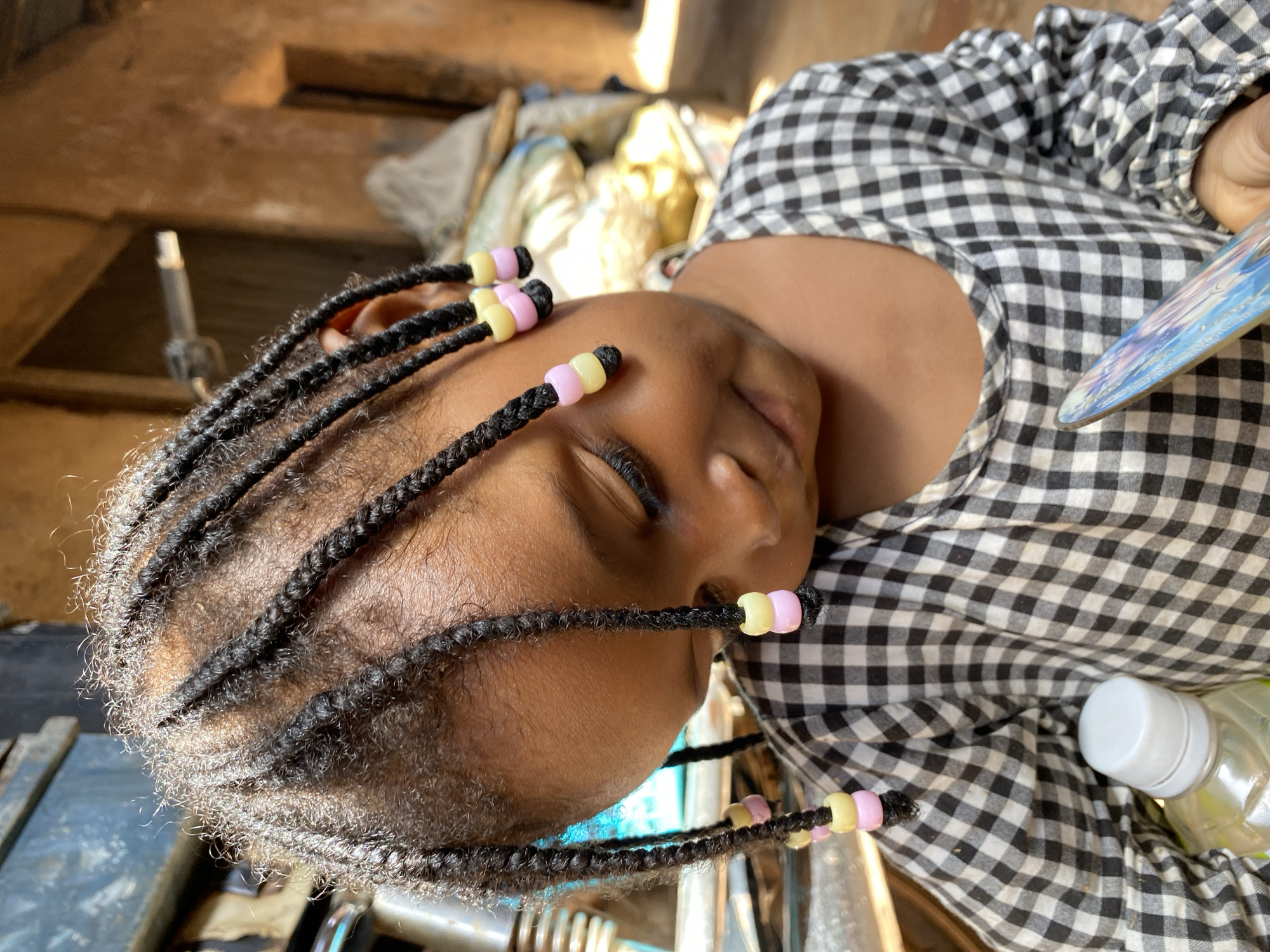 A young girl, with braided hair adorned with pink and yellow beads, rests her head against a checkered fabric. Her eyes are closed, suggesting she is sleeping or resting peacefully. She is wearing a black and white checkered shirt. In the background, blurred objects are visible, including what appears to be a plastic bottle with a white cap. The lighting suggests it might be daytime, with natural light illuminating the scene. The image appears to be taken outdoors, possibly in a rural or less developed setting, given the rough textures and background elements. The overall mood is calm and serene.