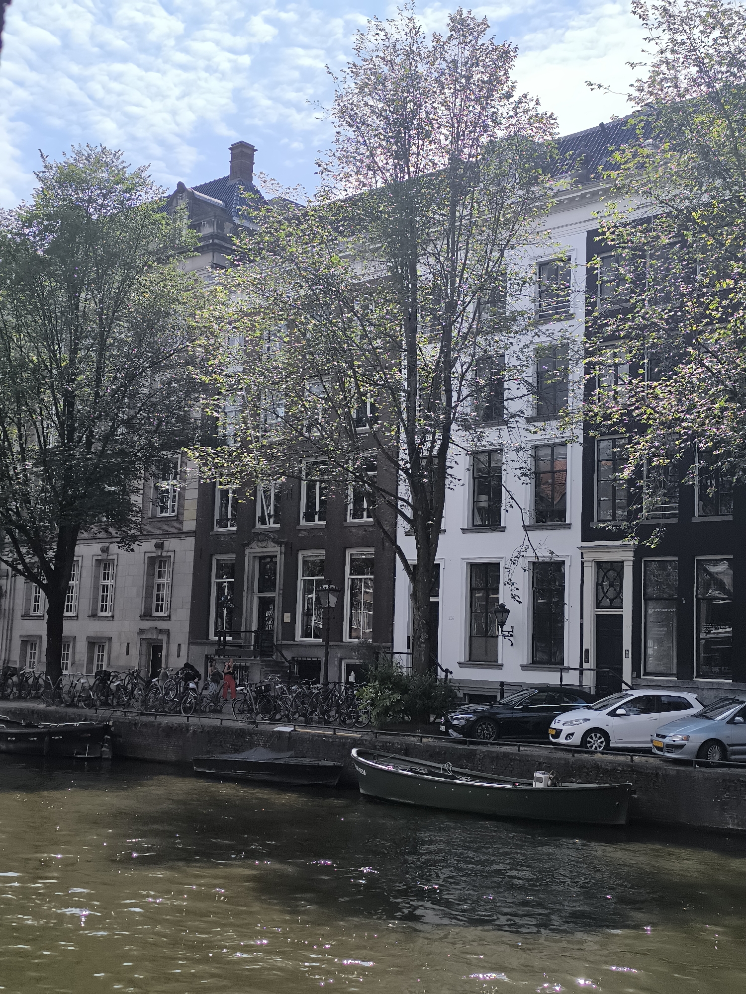 The image depicts a canal-side view of buildings in Amsterdam, Netherlands. The buildings are multi-story, with a mix of architectural styles, including gabled roofs and large windows. Trees with sparse green leaves line the canal, casting dappled shadows on the facades.
In the foreground, the canal water is murky and reflects sunlight, creating shimmering highlights. Several boats are moored along the canal edge, including a long, dark green boat in the middle ground and other smaller vessels.
Along the embankment behind the canal, a row of bicycles is parked. A few people are visible near the bicycles, including one person in a red outfit. Cars are parked along the street behind the embankment, including a black sedan, a white hatchback, and a silver sedan. The sky above is partly cloudy. The overall scene suggests a typical urban canal setting in Amsterdam.