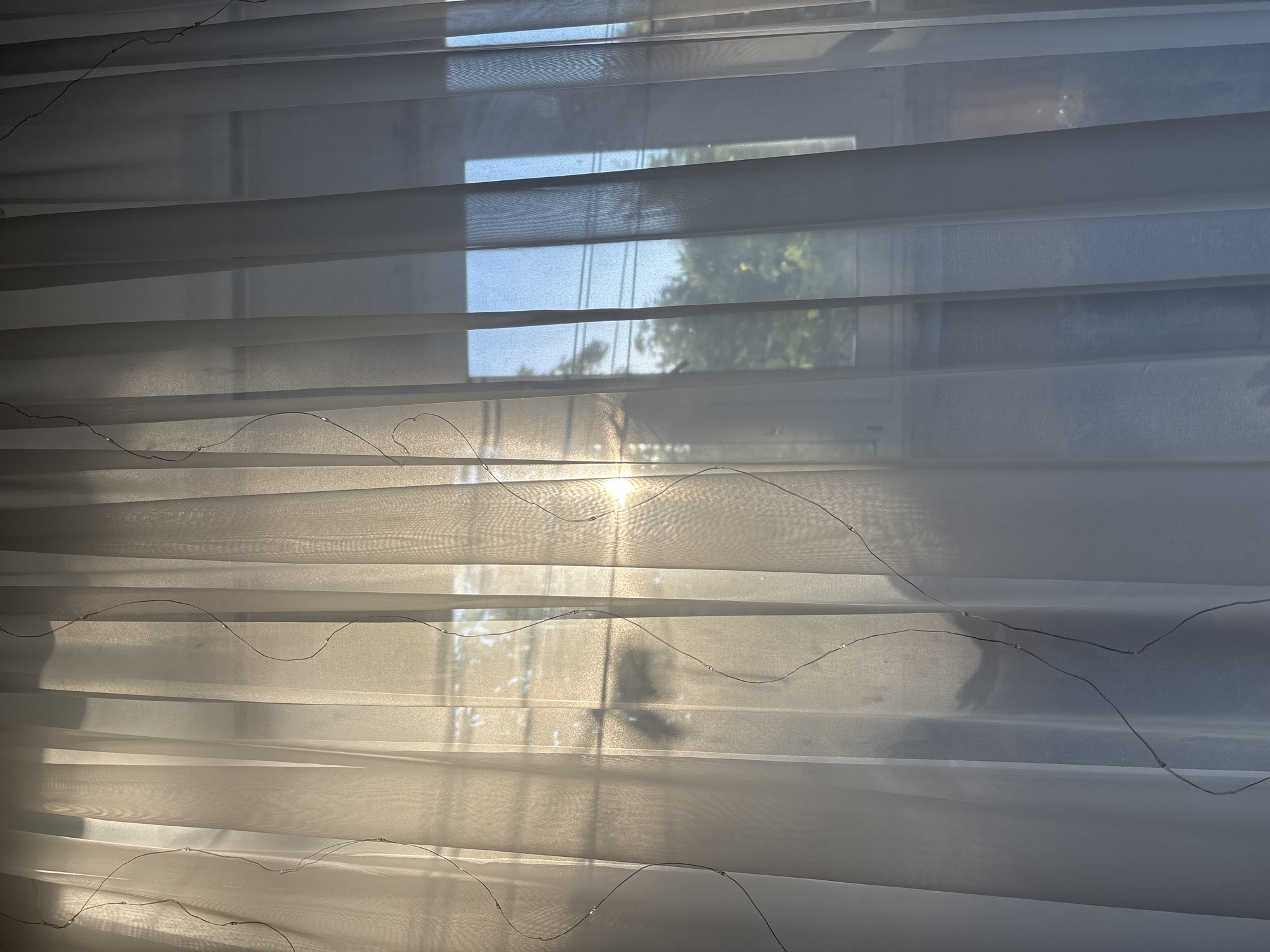The image shows a close-up of sheer horizontal blinds against a window. The blinds are made of a sheer, translucent fabric, creating a layered effect. Thin, delicate wires with small lights are strung across the fabric of the blinds, creating subtle, decorative patterns.
Through the blinds, a window is visible. The window is divided into horizontal panes by the blinds. Sunlight is shining through the window, creating bright highlights and casting soft shadows. The sunlight is diffused by the sheer blinds, giving the scene a warm and gentle glow.
Visible through the window is an outdoor scene with trees and what appear to be telephone or power lines against a bright blue sky. The time of day appears to be morning or late afternoon, based on the angle and intensity of the sunlight. The overall mood of the image is serene and peaceful, with a soft, ethereal quality.
There is no visible text in the image.