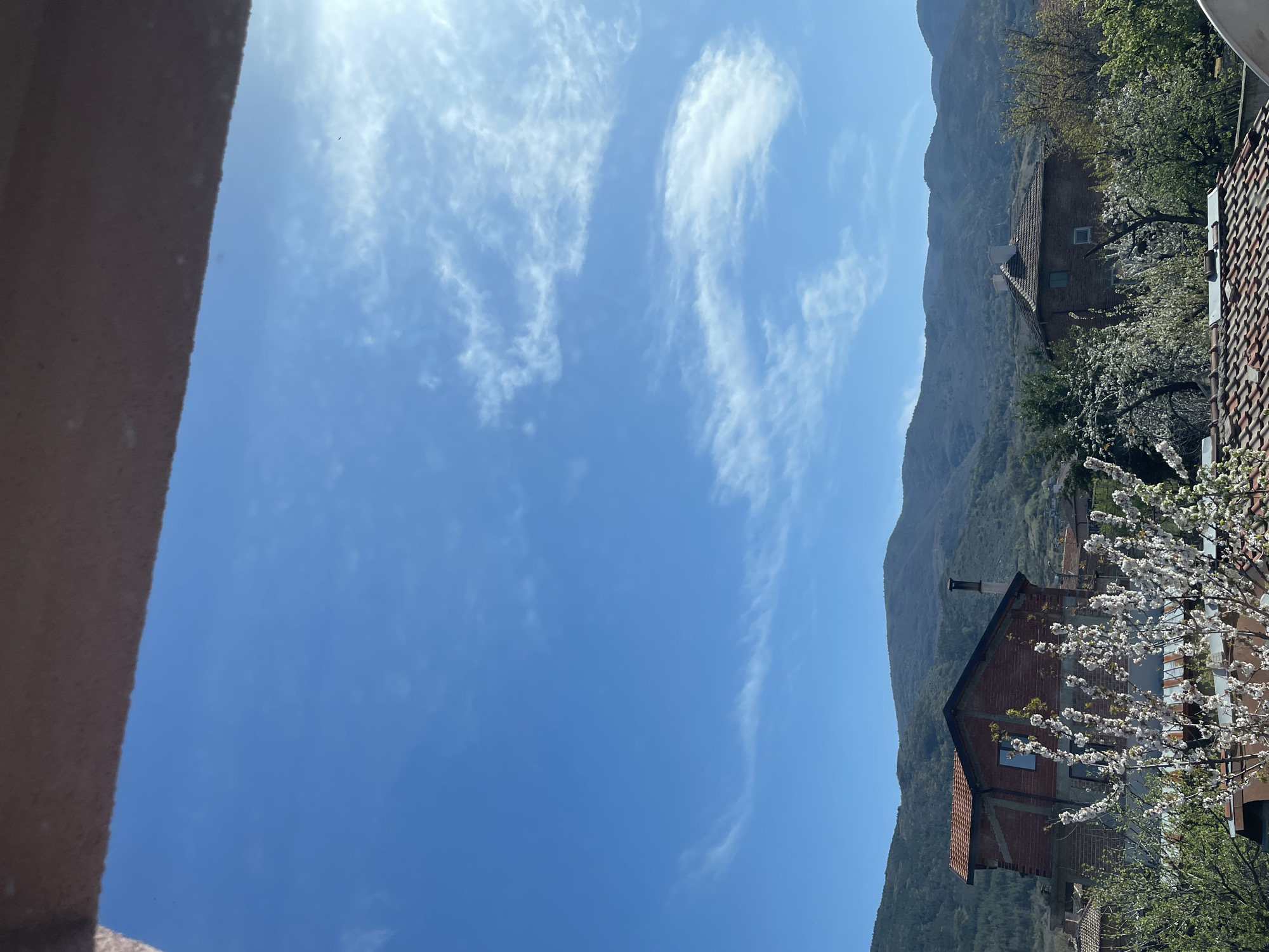 The image presents a view towards a partially clear, bright blue sky with light wispy clouds. On the right side of the frame, there's a mountainous landscape with sparse trees and vegetation. Within this landscape, several buildings are visible, including houses with red-tiled roofs, partially obscured by flowering trees or bushes. The framing is slightly off-kilter, giving a tilted perspective. The overall impression is of a rural or semi-rural setting on a clear, sunny day.