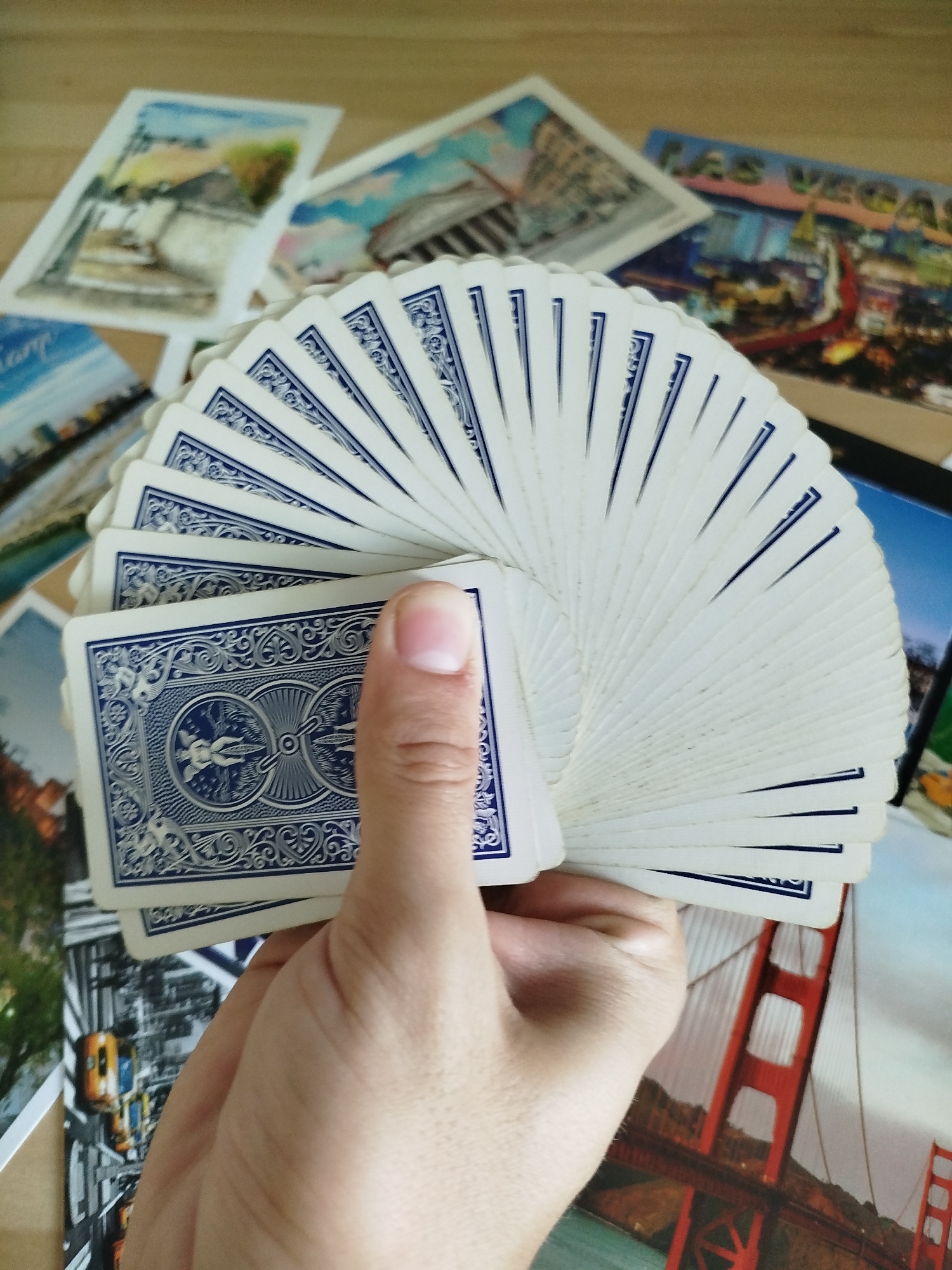 In the foreground, a person's hand holds a fanned-out deck of playing cards. The cards have a blue and white patterned back. Behind the cards, several postcards are visible, some of which appear to be travel-themed, depicting scenes such as buildings, cityscapes, and a suspension bridge. The background is a wooden surface. There is no indication of the city of Varna, Bulgaria, in the image.
