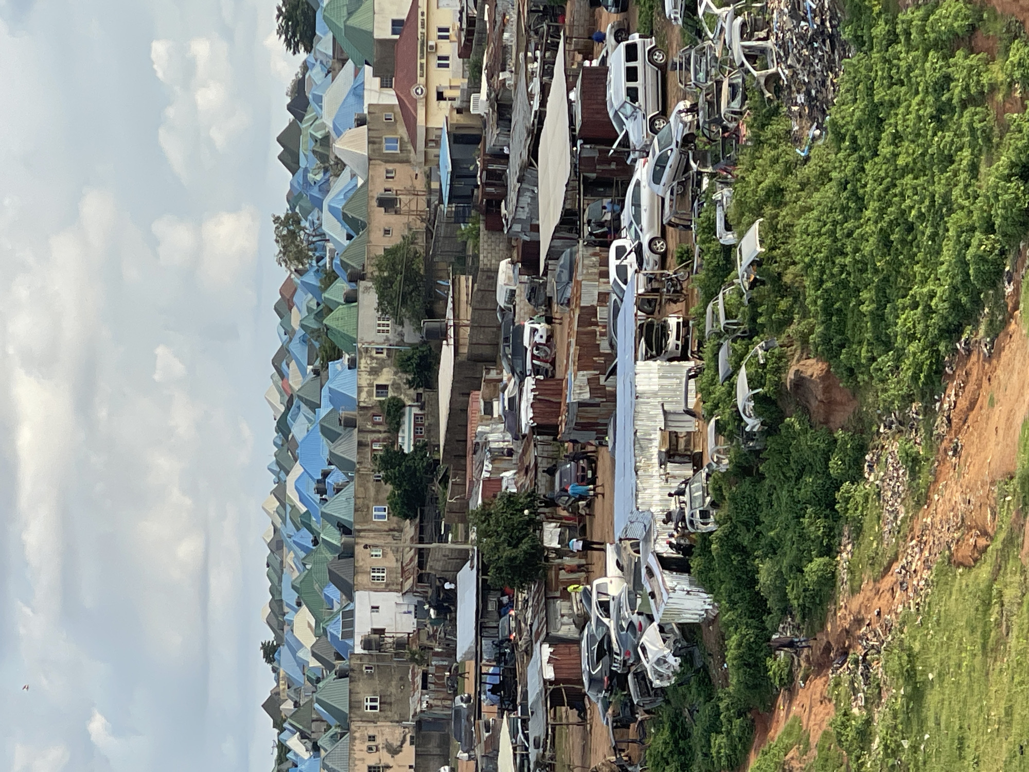 This is an aerial shot of a densely populated area in Abuja, Nigeria, characterized by a sprawling collection of residential buildings and an informal auto repair and parts market. The residential buildings feature distinct cone-shaped roofs in various shades of blue, green, and off-white, clustered tightly together. Below this residential area, the scene transitions to a more industrial and chaotic environment.
The foreground and middle ground are dominated by an extensive junkyard or car repair lot. Numerous vehicles, many of them dismantled or in various states of disrepair, are scattered across the landscape. Some are complete cars, while others are merely chassis or stacks of car parts. Various structures, likely makeshift workshops or storage areas, constructed from corrugated metal and wood, are interspersed among the vehicles.
Several individuals are visible in the scene, engaged in various activities. Some appear to be working on the vehicles, while others are standing around or walking through the area. The ground in the lower part of the image is a mix of dirt paths and green vegetation, with scattered debris and rubbish visible in some areas. The overall impression is one of a bustling, somewhat unorganized urban environment where residential and commercial activities, specifically auto salvage and repair, are interwoven.