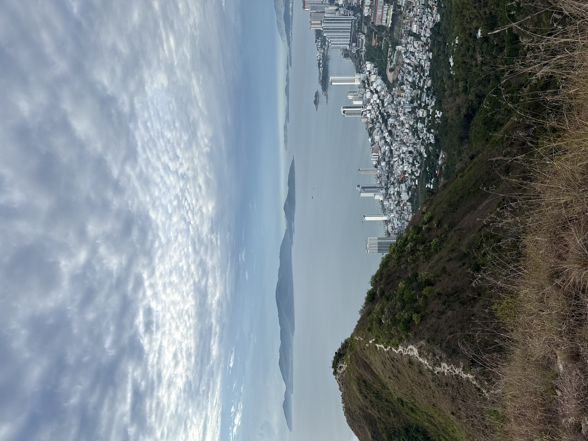 The image presents a panoramic view of Bac Nha Trang Ward, Vietnam. The foreground features a hillside with a winding trail and sparse vegetation. The cityscape is visible along the coast, consisting of dense clusters of buildings and high-rise structures. A body of water extends into the distance, separating the city from a series of mountainous islands under a sky with cloudy cover.