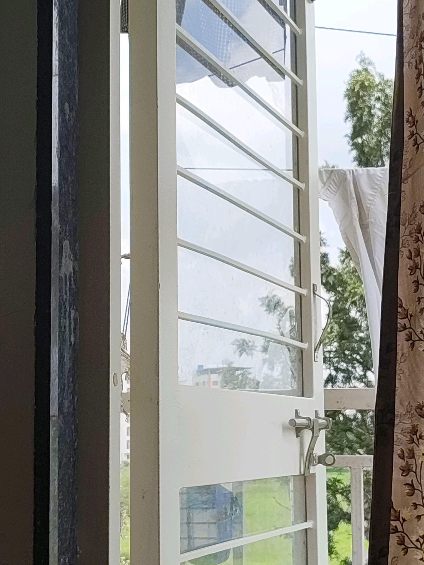The image shows an open white window with horizontal bars, revealing a green outdoor space with trees and a white cloth hanging. A metal latch is visible on the window. A patterned curtain is on the right side of the frame, and a dark vertical object, possibly a column or wall, is on the left. The background suggests an urban or semi-urban environment. The scene is captured from an interior perspective looking outwards in Tathawade, India.