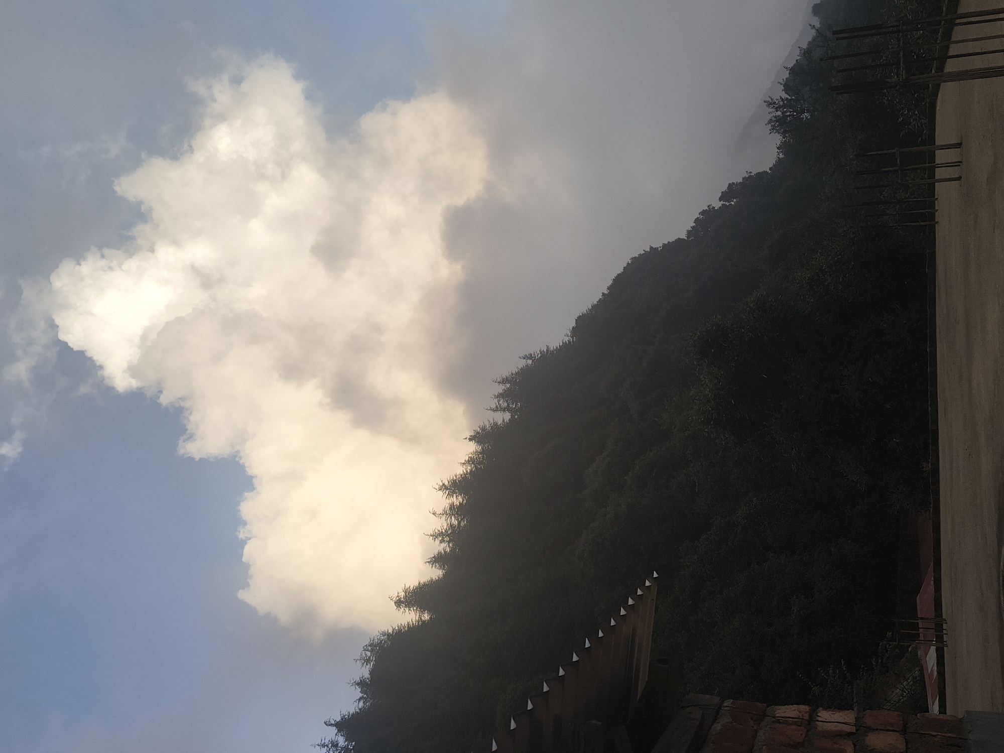 A photograph taken from a rooftop in Dharamkot, India, captures a dramatic sky scene. The sky is a mix of blue and overcast with large, luminous clouds that appear to be lit from behind, creating a soft, glowing effect. A heavily forested hillside, densely packed with evergreen trees, slopes diagonally across the frame from the top left towards the bottom right. The trees are dark silhouettes against the brighter sky. In the foreground, the edge of a building's roof is visible on the left, constructed with corrugated metal and brickwork. The rooftop surface is a light brown concrete. In the mid-ground and background, more indistinct structures and bare vertical supports suggest ongoing construction or an unfinished building. The overall atmosphere is moody and serene, with the natural elements of the sky and forest dominating the scene.